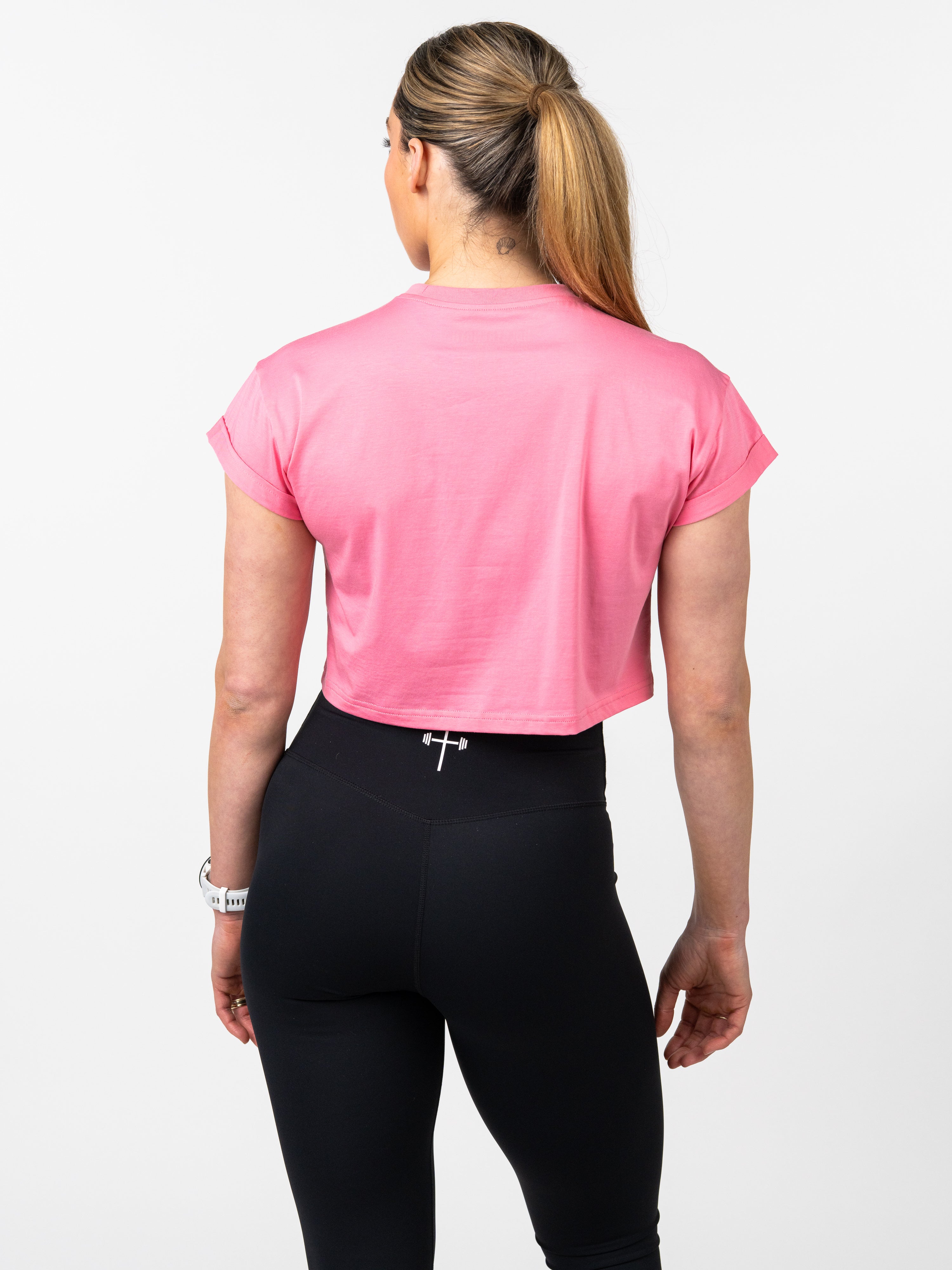 Woman wearing pink Cross Essential Crop Tee and black leggings with subtle Christian cross and barbell design on the waistband, shown from the back in a studio setting.