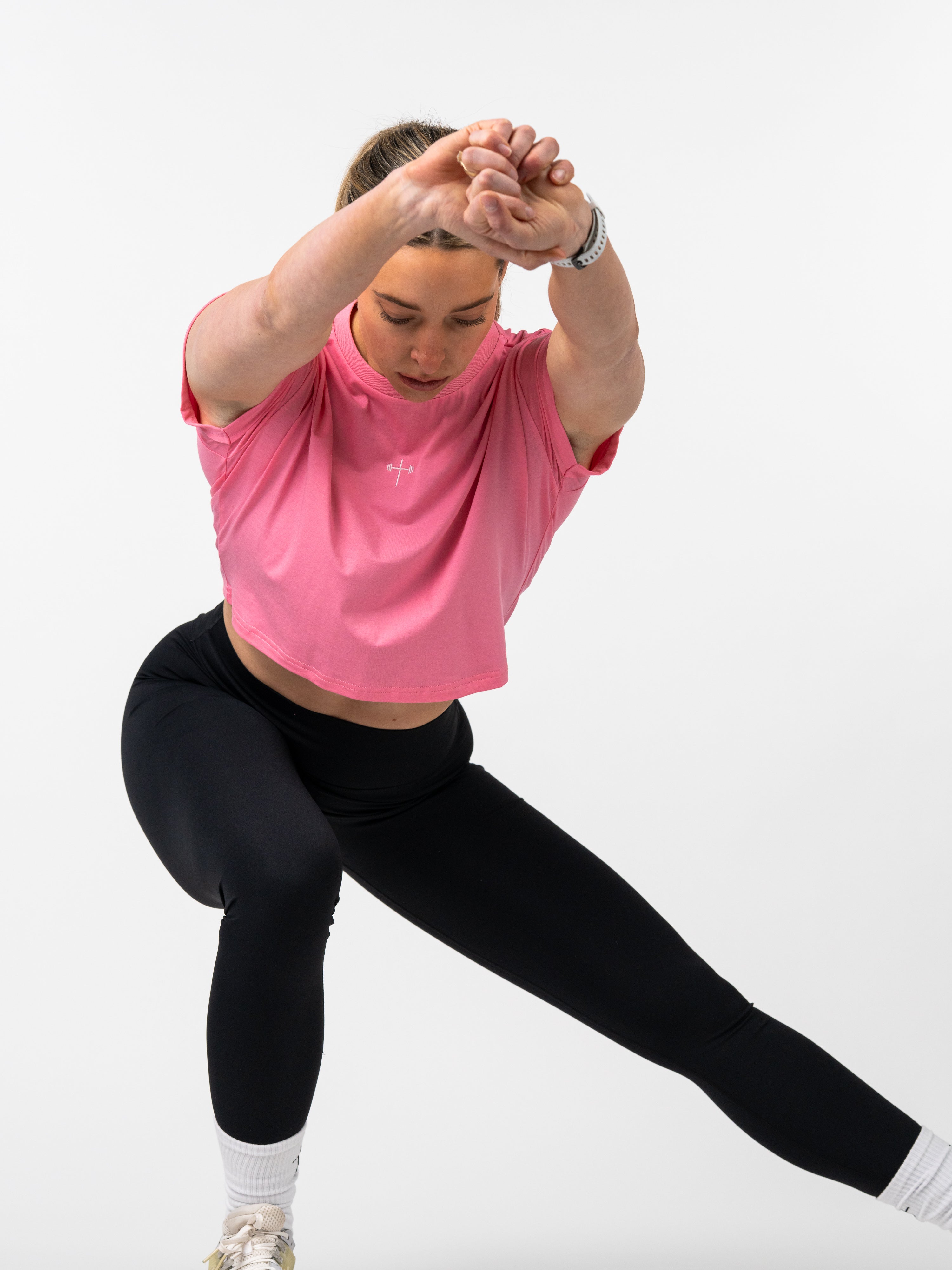 Woman in black leggings performing side lunge stretch while wearing a pink Cross Essential Crop Tee with a subtle cross and barbell design, showcasing Christian fitness apparel.