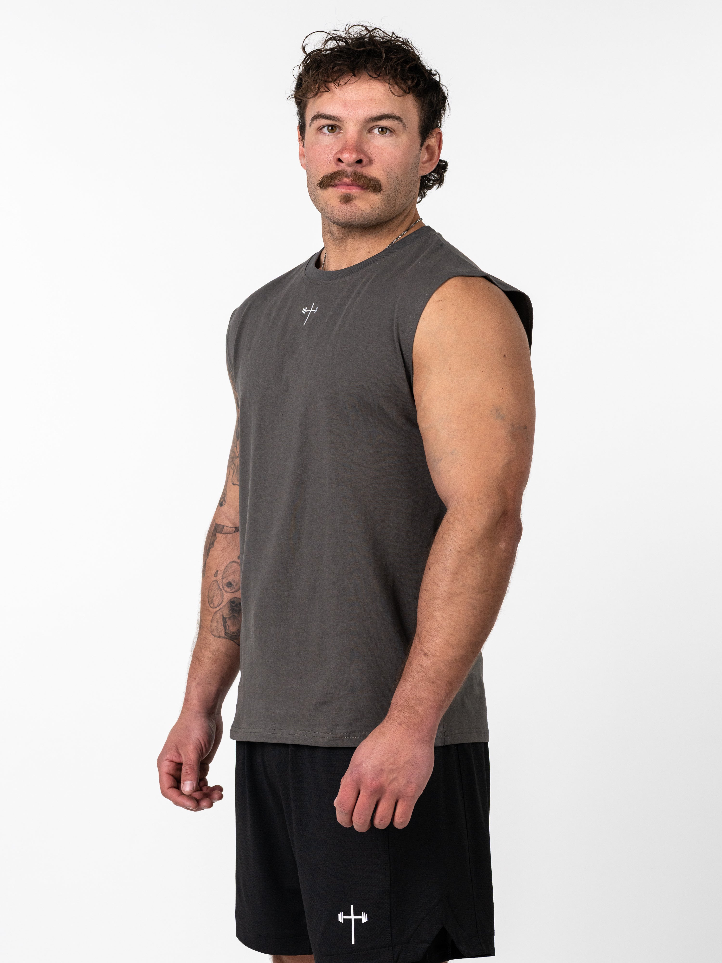 Man wearing a charcoal Scripture Premium Sleeveless Tank with a small cross and barbell symbol on the chest, paired with black shorts featuring a similar logo, posing against a white background.