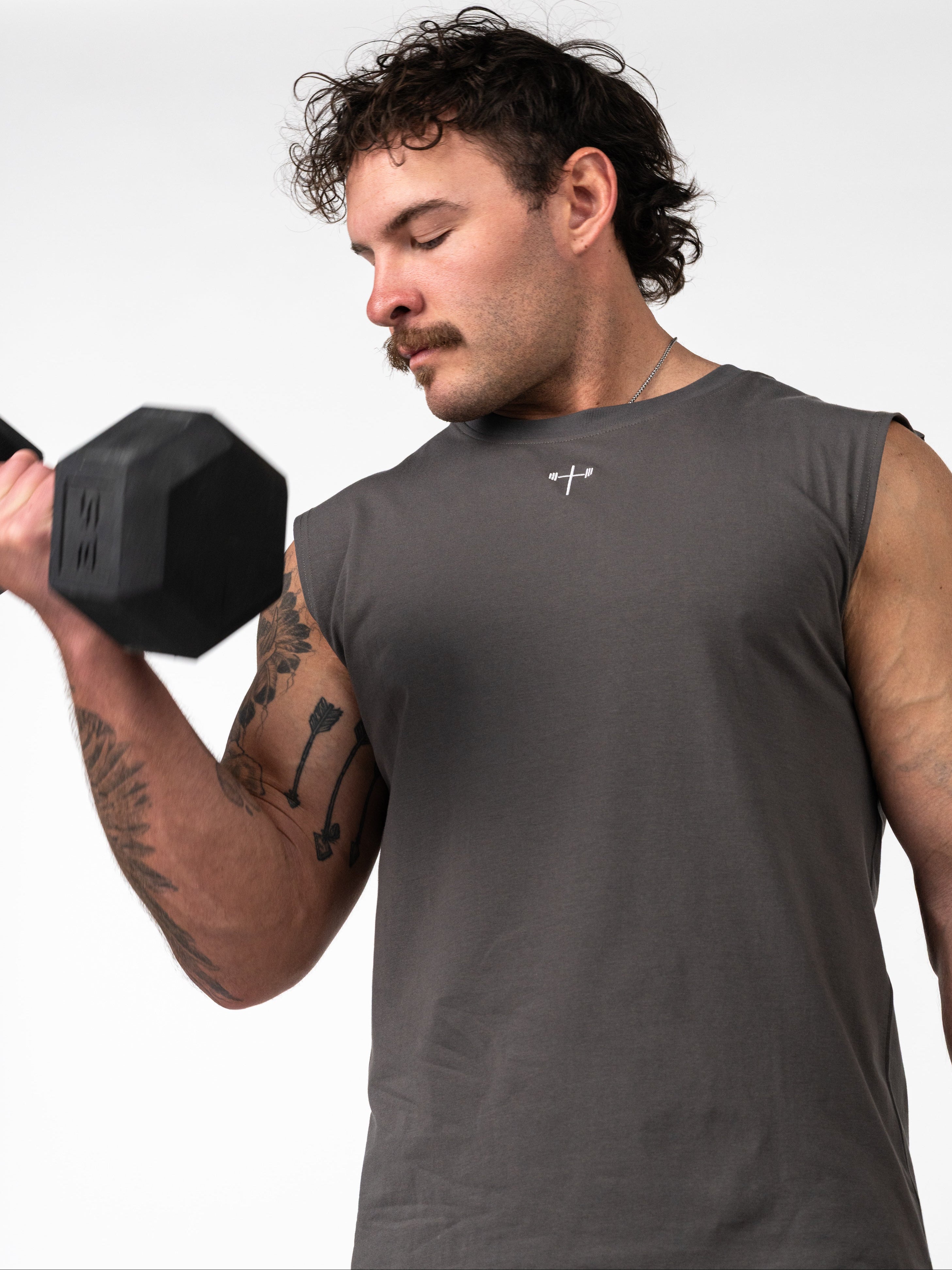 Man lifting a dumbbell while wearing a dark gray Scripture Premium Sleeveless Tank featuring a small Christian cross with a barbell design on the chest.