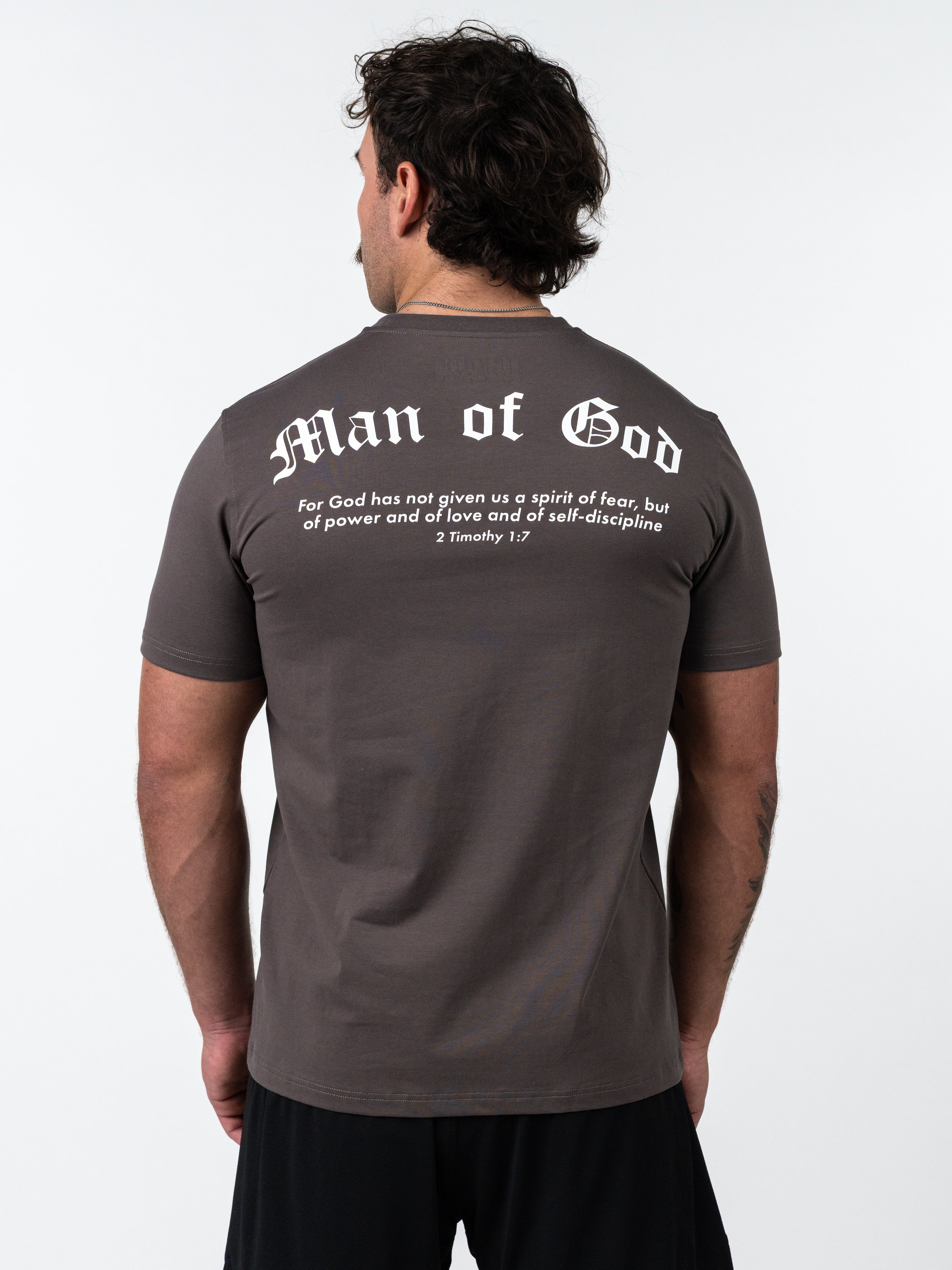 Man wearing a Kingdom Performance Tee in iron grey with white "Man of God" text and Bible verse on the back, standing facing away against a plain background.