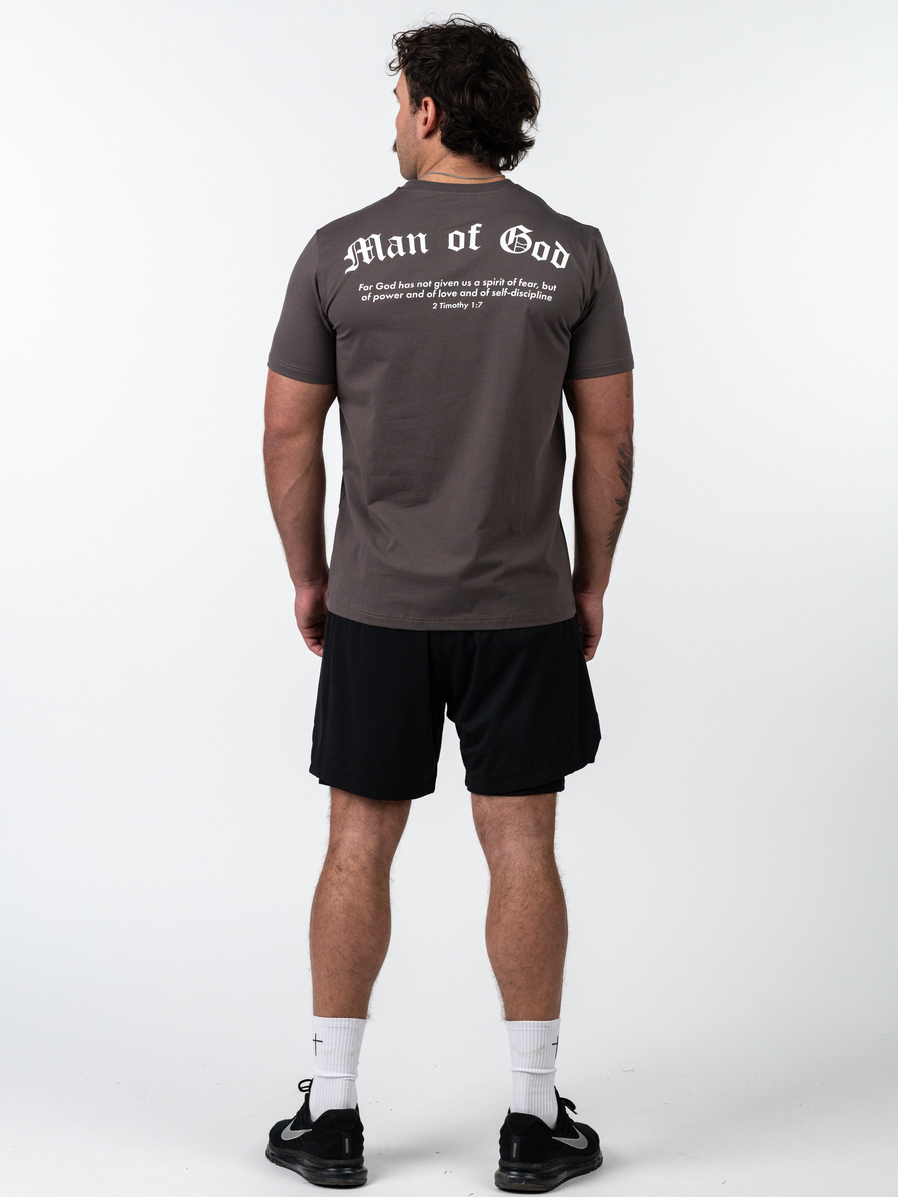 Man wearing a Kingdom Performance Tee in dark grey with "Man of God" text and Bible verse on the back, paired with black athletic shorts, white socks, and black shoes.