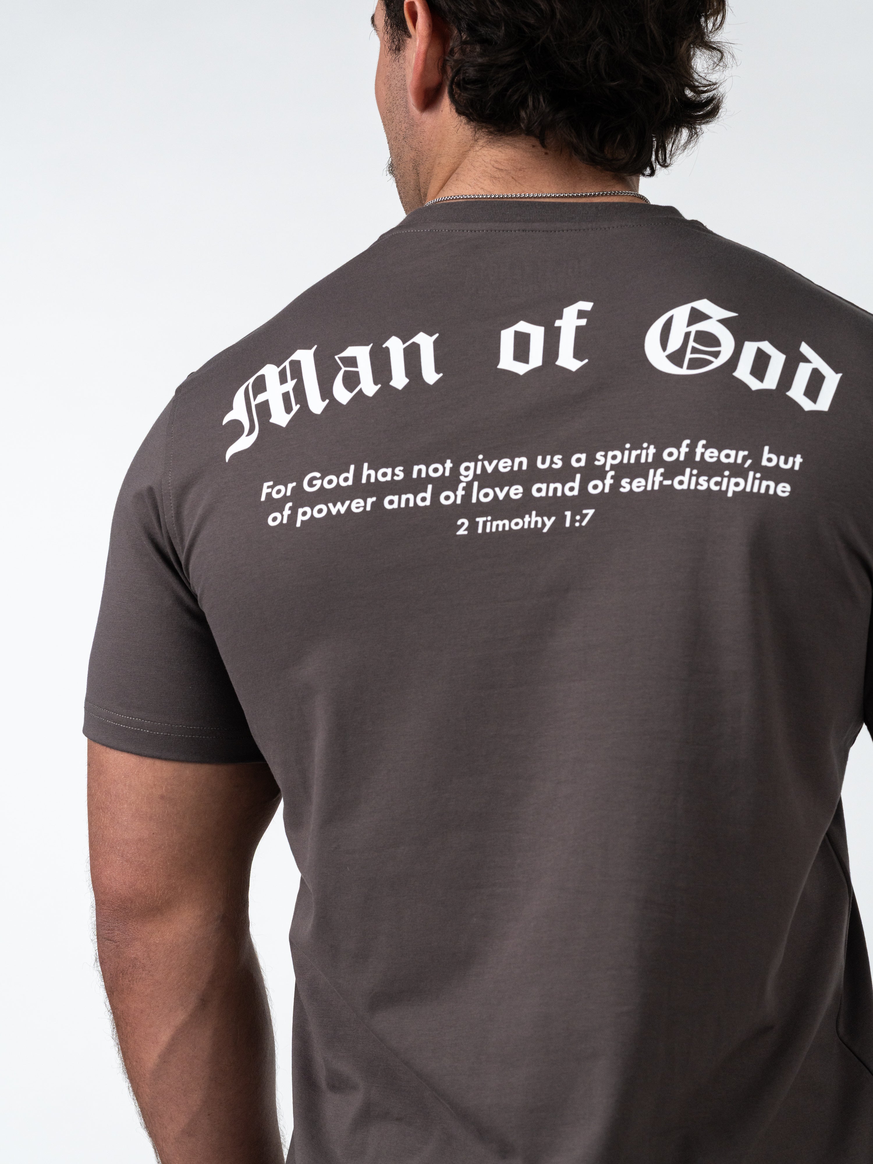 Man wearing an Iron Grey Kingdom Performance Tee showing the back design with "Man of God" text and a Bible verse from 2 Timothy 1:7 in white, Christian gym clothing style.