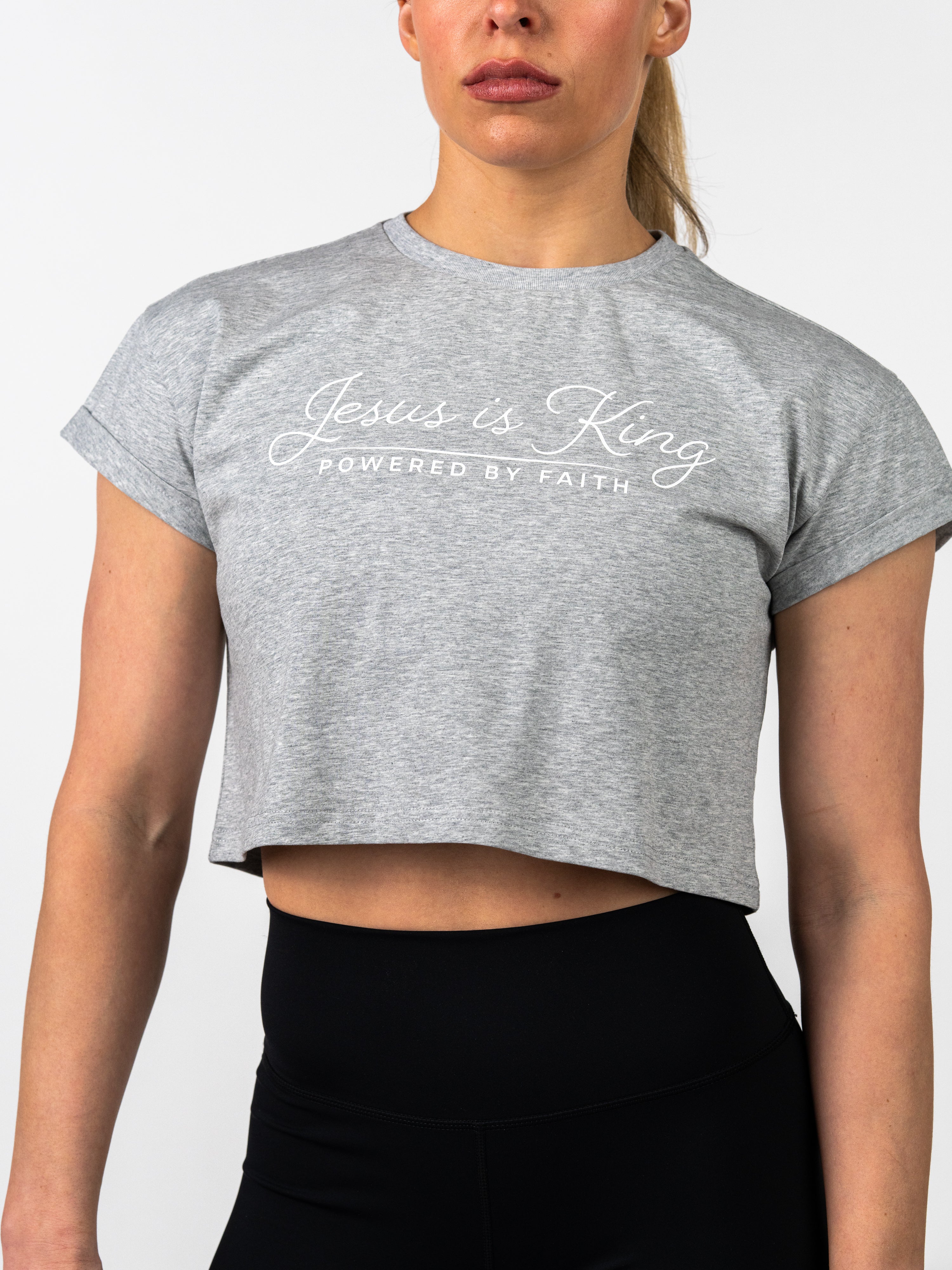 Woman wearing a gray Jesus is King Essential Crop Tee with white script text, paired with black high-waisted athletic leggings, showcasing Christian fitness apparel.