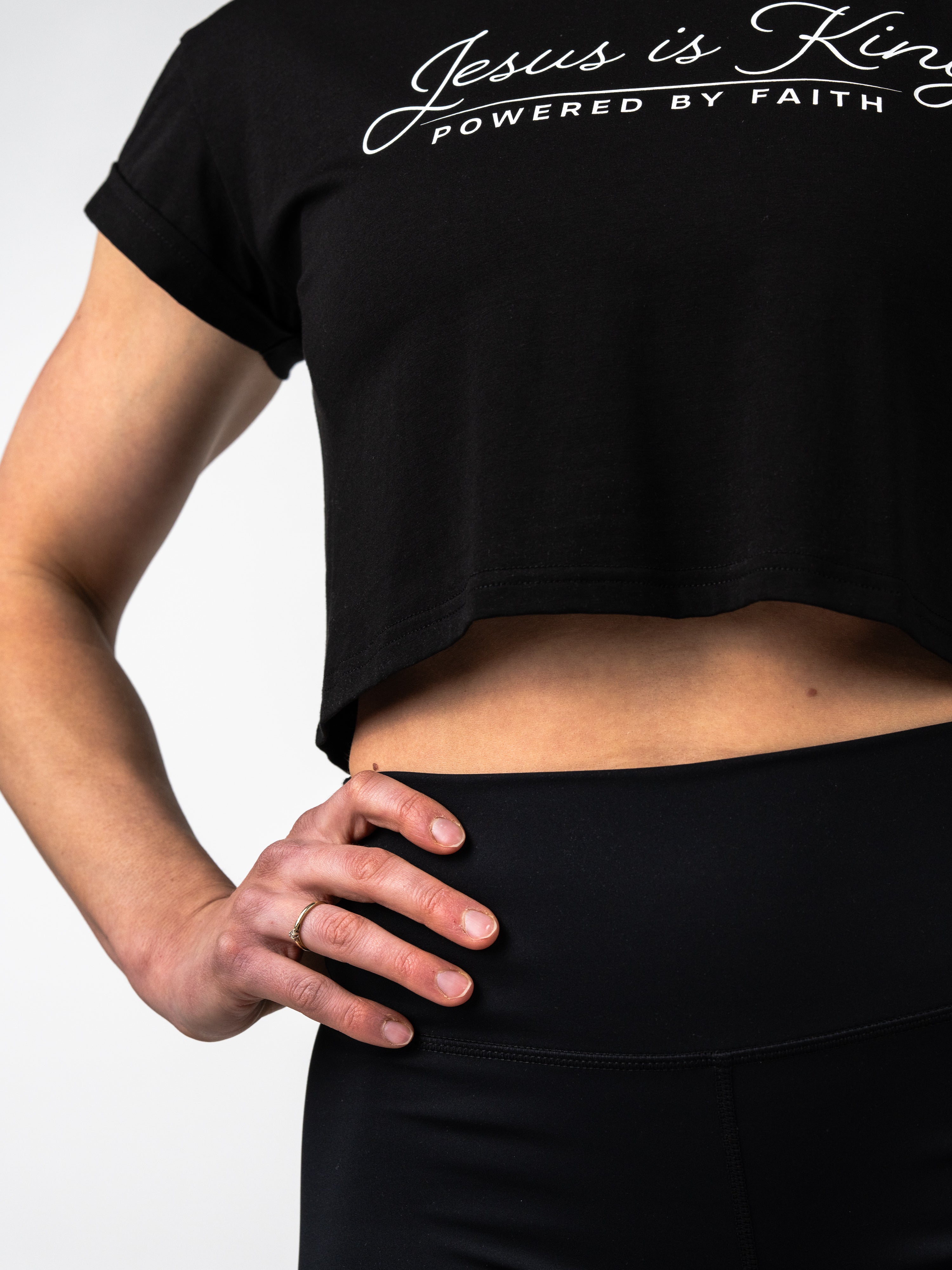Close-up of a person wearing a black Jesus is King Essential Crop Tee with white text and high-waisted black athletic leggings, showcasing Christian fitness apparel.