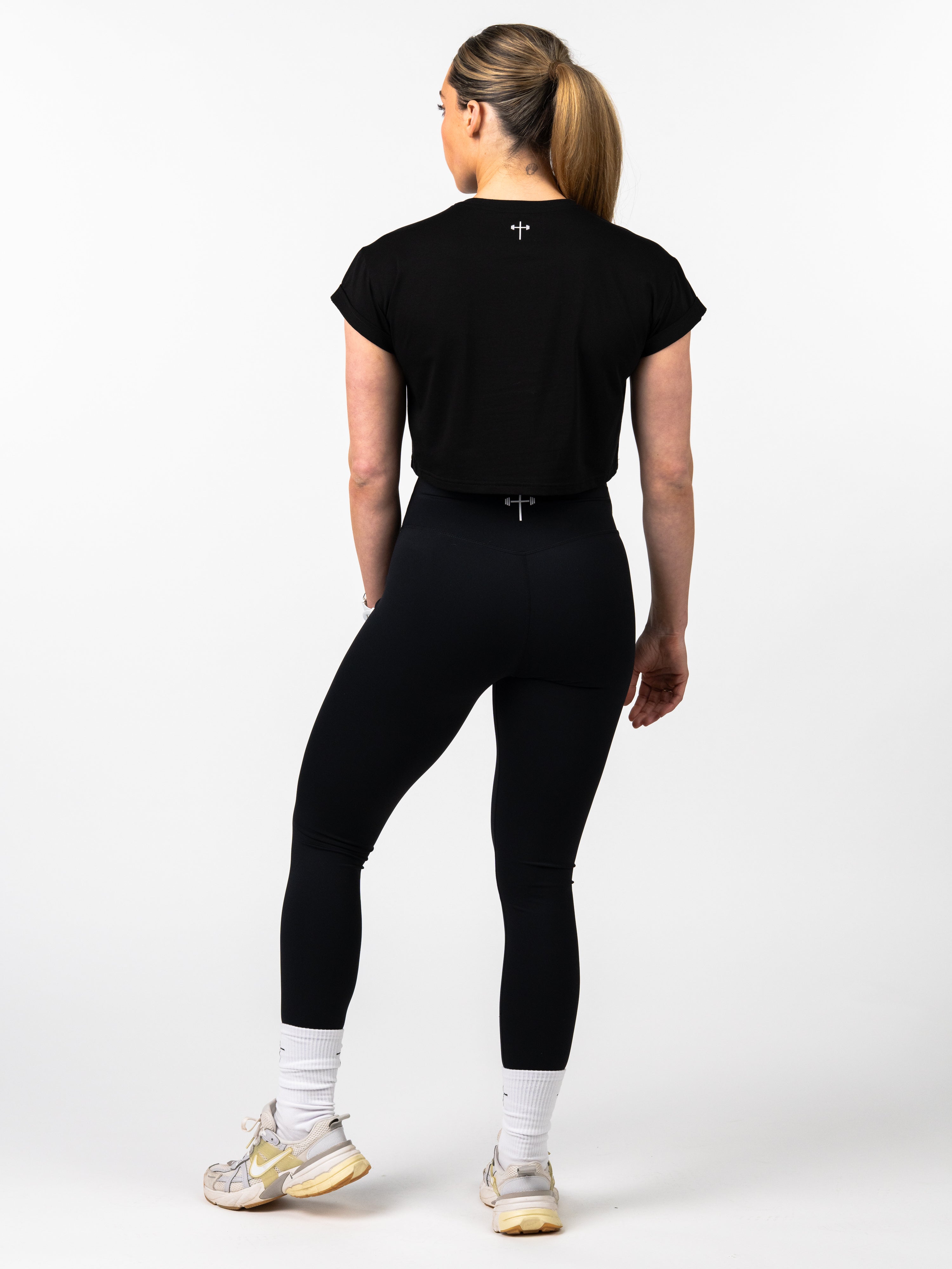Woman shown from the back wearing a black Jesus is King Essential Crop Tee with a small cross detail near the neckline, paired with black leggings and white athletic shoes.