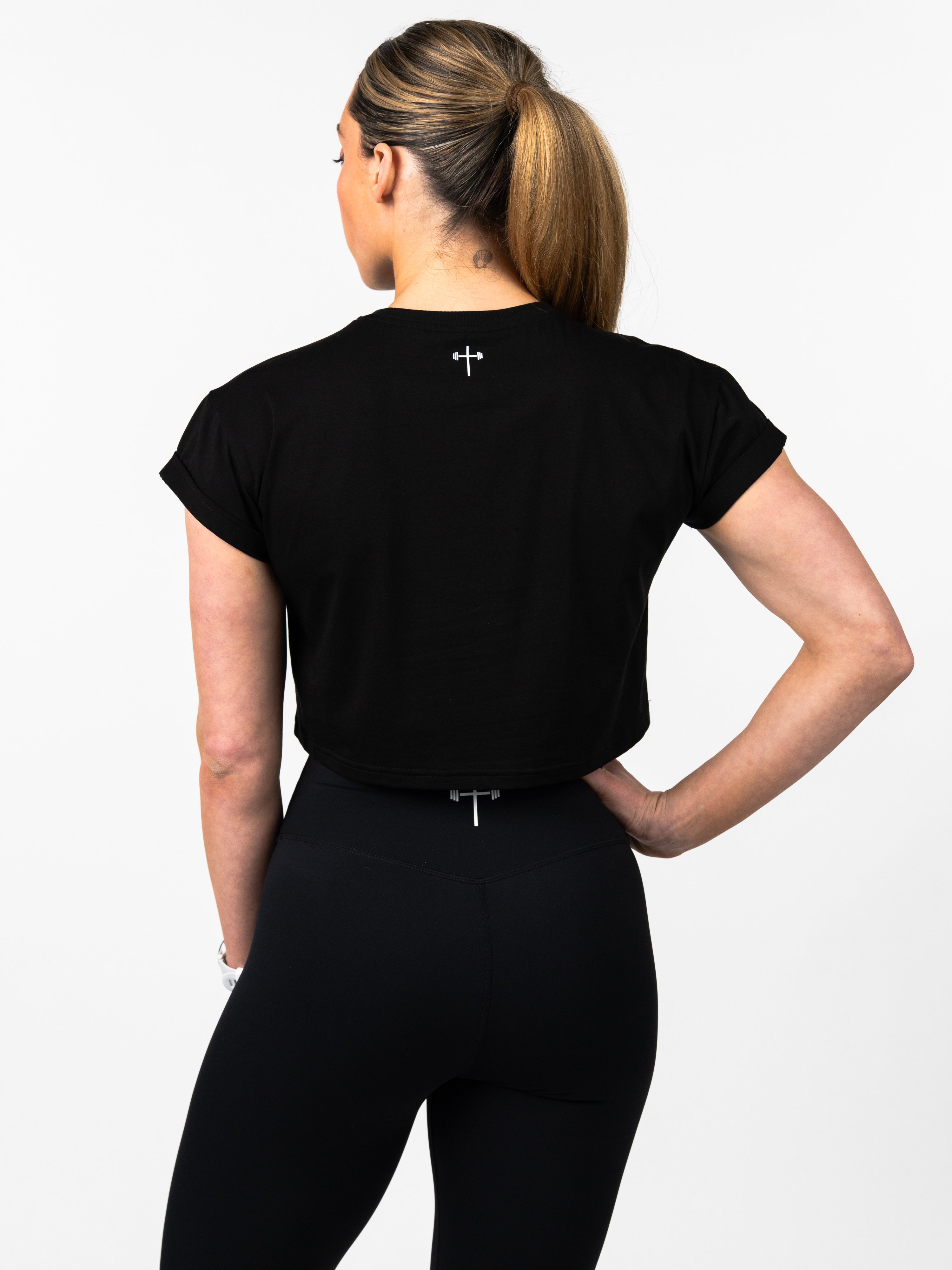 Woman wearing a black Jesus is King Essential Crop Tee from the back, featuring a small white cross design near the neckline, paired with black workout leggings.