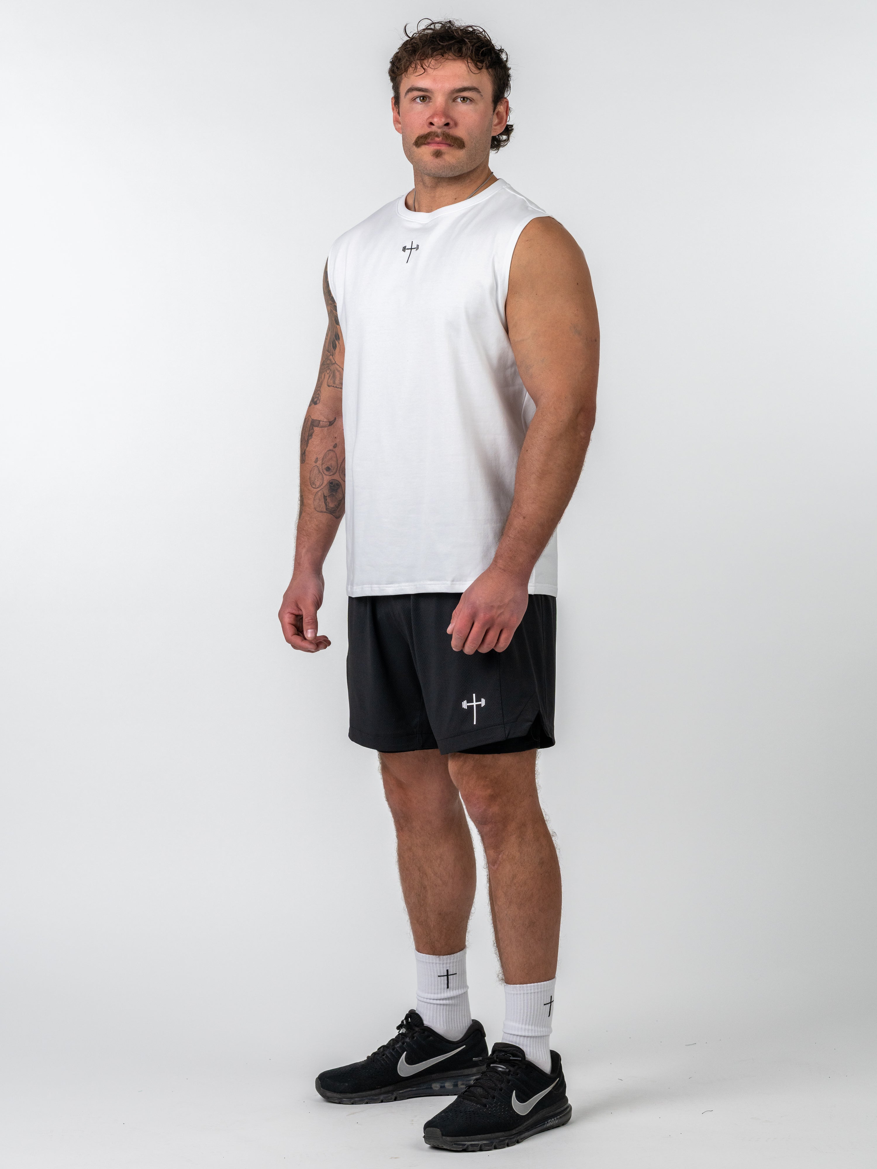 Man wearing a white Sacred Heart Premium Sleeveless Tank paired with black athletic shorts, white socks with a cross design, and black Nike running shoes, standing against a plain background.