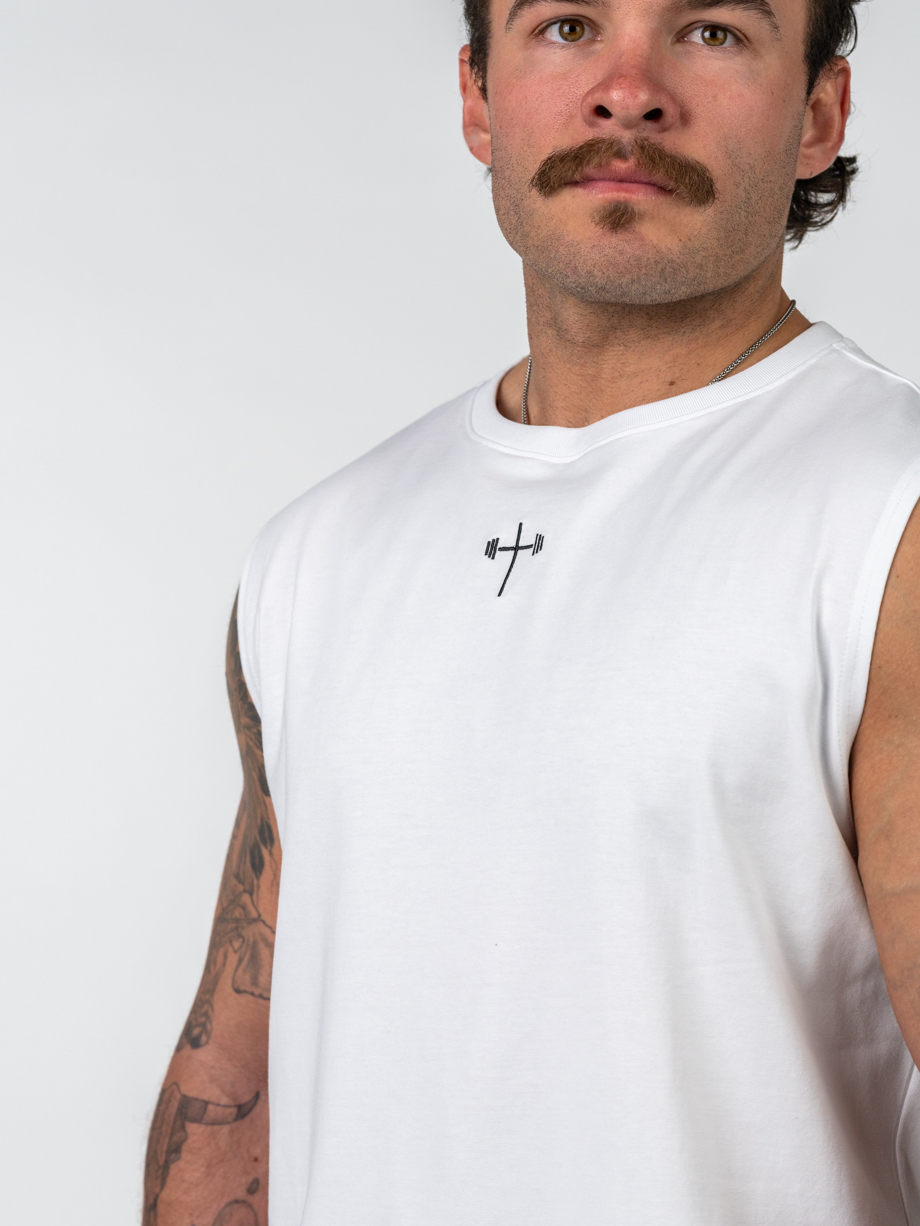 Man wearing a white Sacred Heart Premium Sleeveless Tank featuring a small black barbell cross symbol on the chest, representing Christian fitness apparel and faith-based workout clothing.