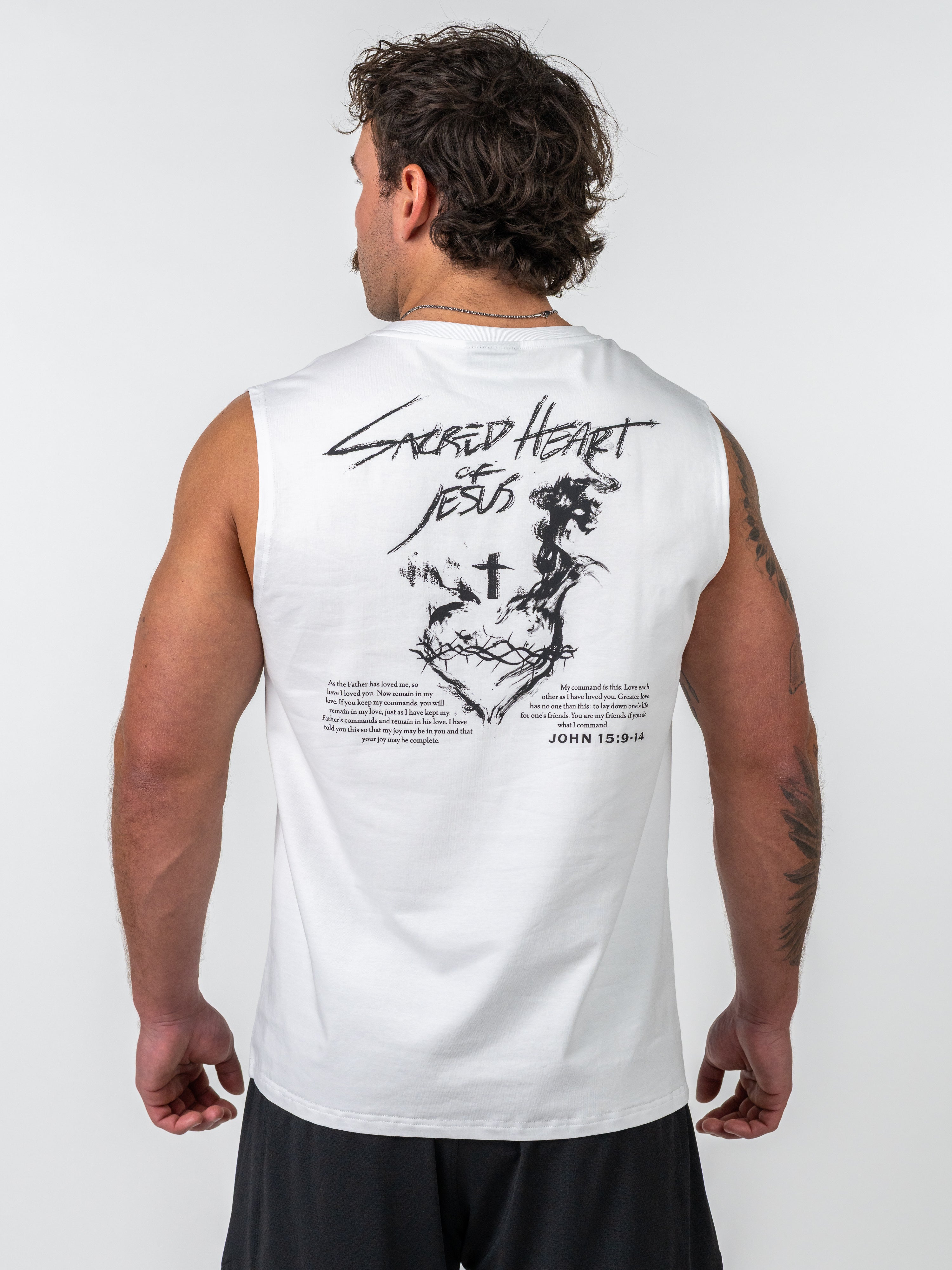 Man wearing a white Sacred Heart Premium Sleeveless Tank showing the back with black graphic artwork of a cross above a crown of thorns and scripture text from John 15:9-14, representing Christian fitness apparel.