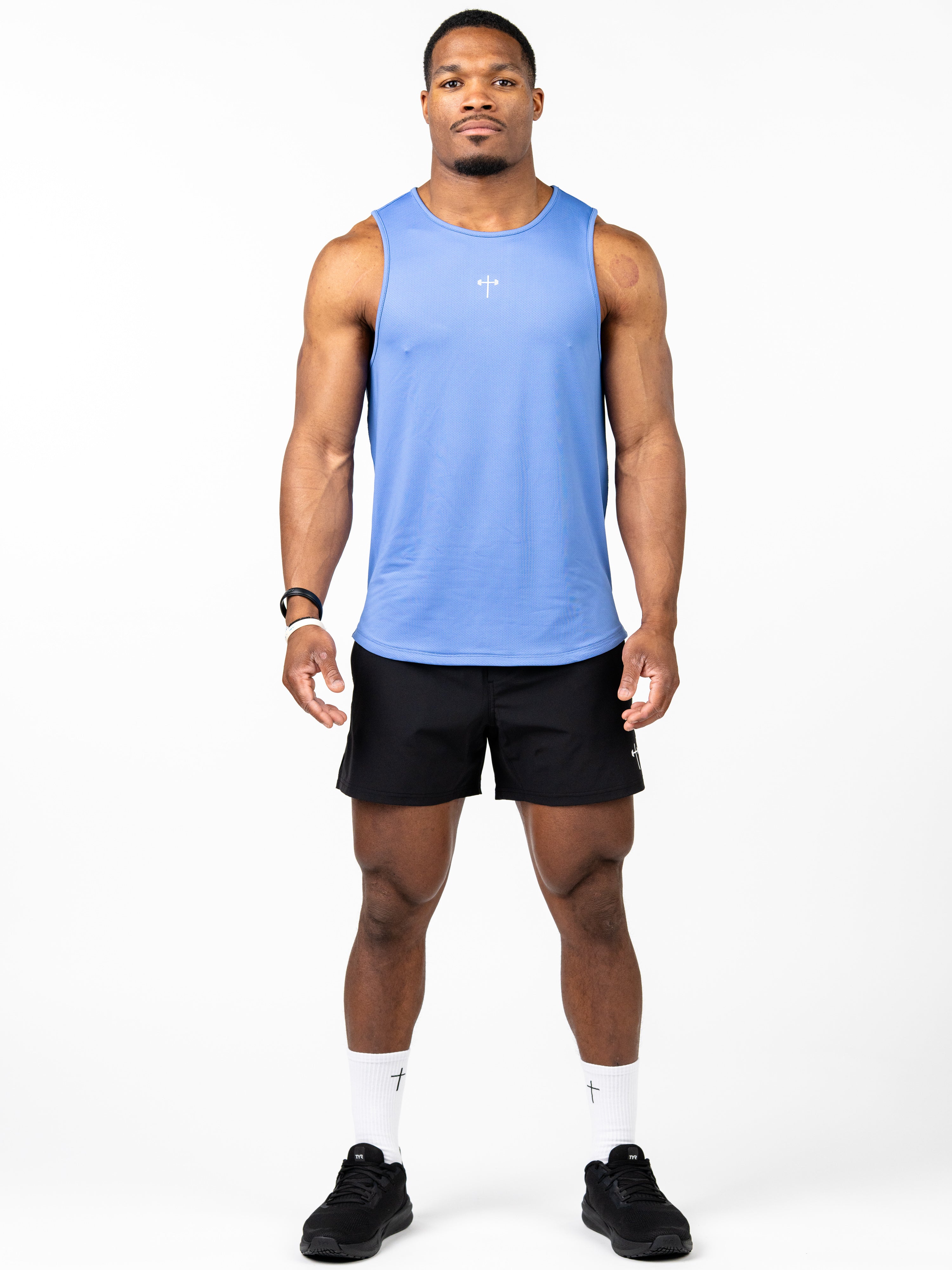 Athletic man wearing a blue Signature Tank with a small Christian cross and barbell design on the chest, paired with black shorts and white socks featuring a cross logo.