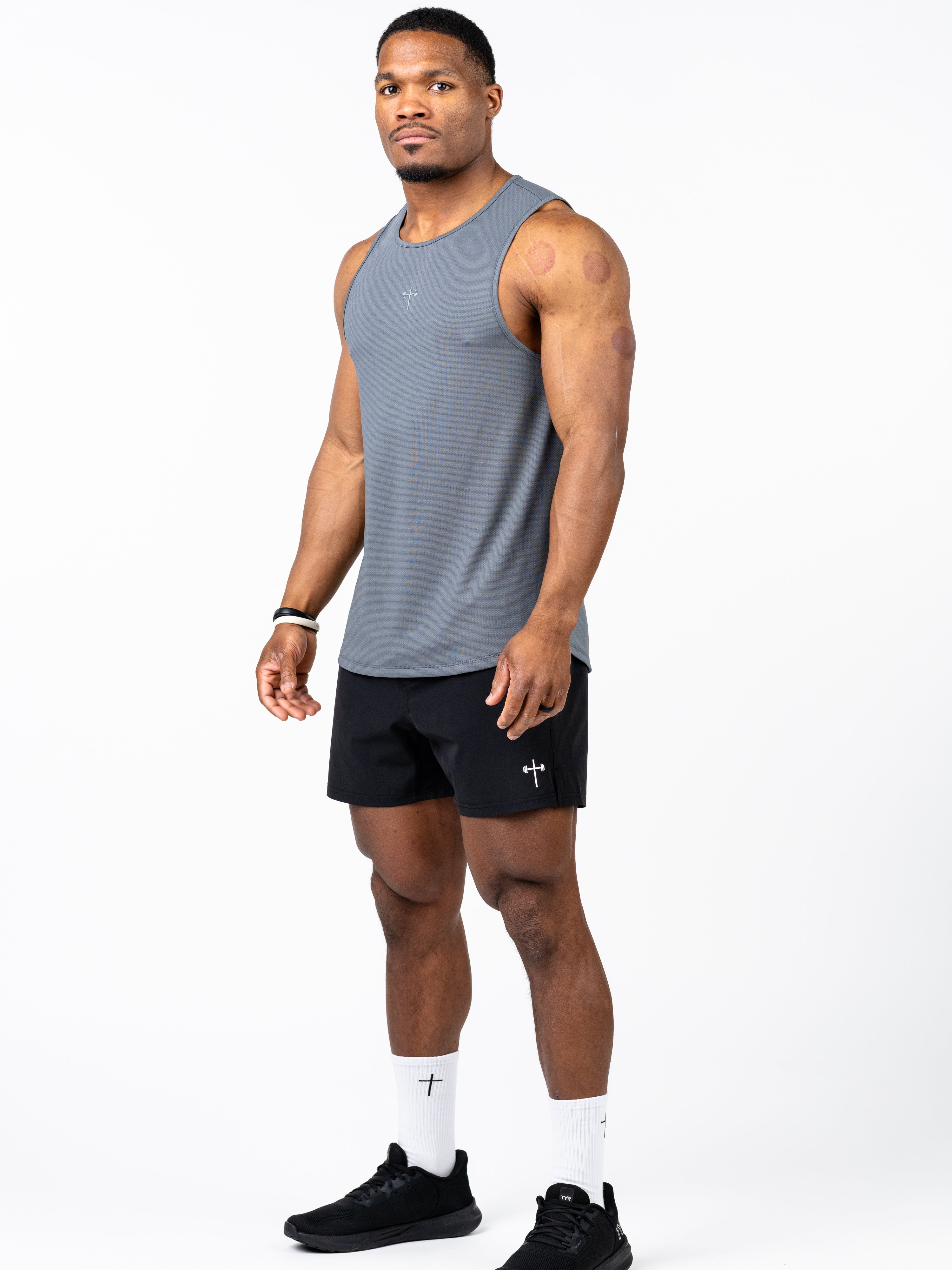 Athletic man wearing a gray Signature Tank designed for performance and durability, paired with black shorts featuring a subtle Christian cross logo, white socks, and black athletic shoes.