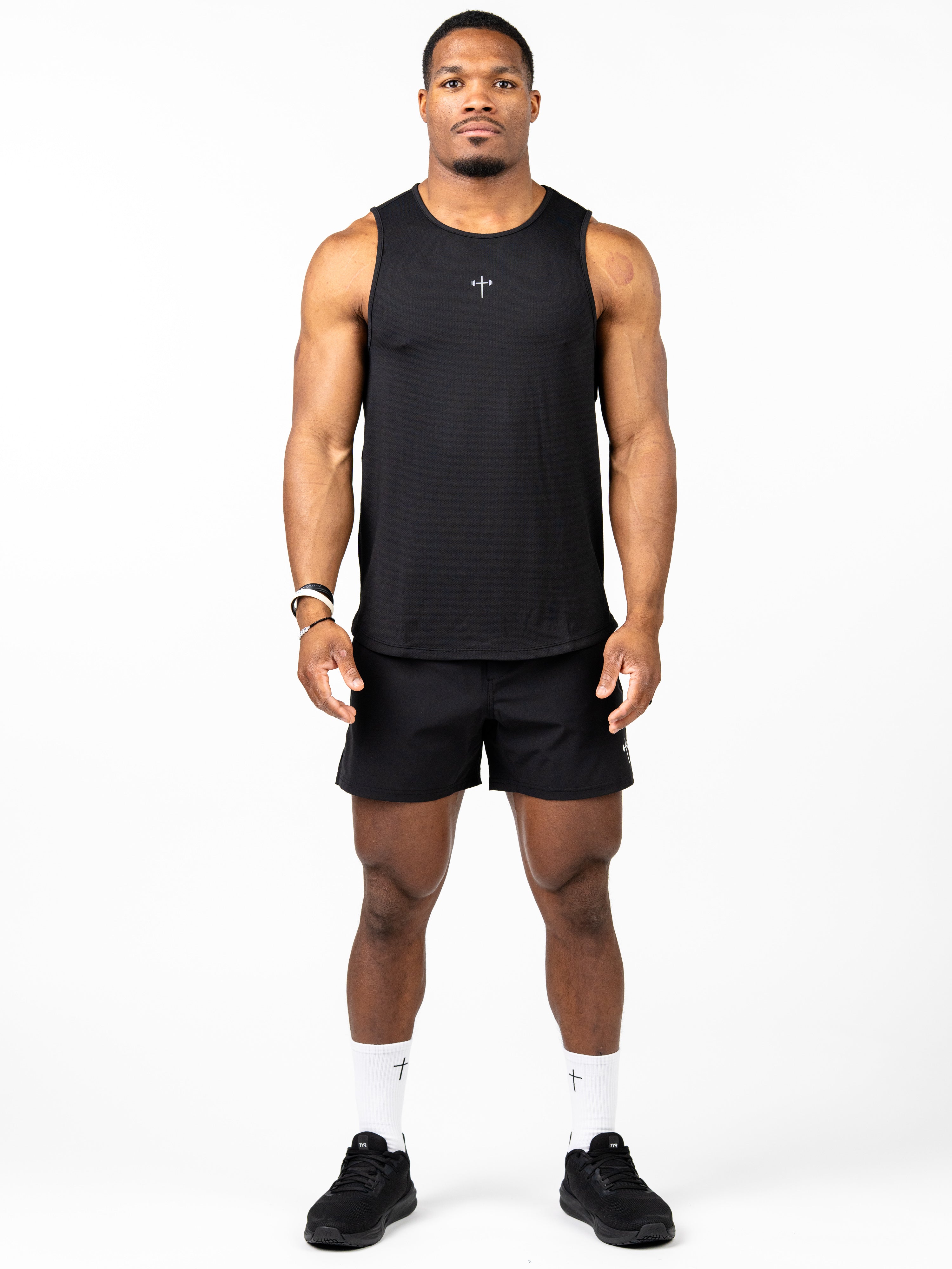 Man wearing a black Signature Tank with a small cross design on the chest, paired with black shorts, white socks featuring a cross, and black athletic shoes, standing against a white background.