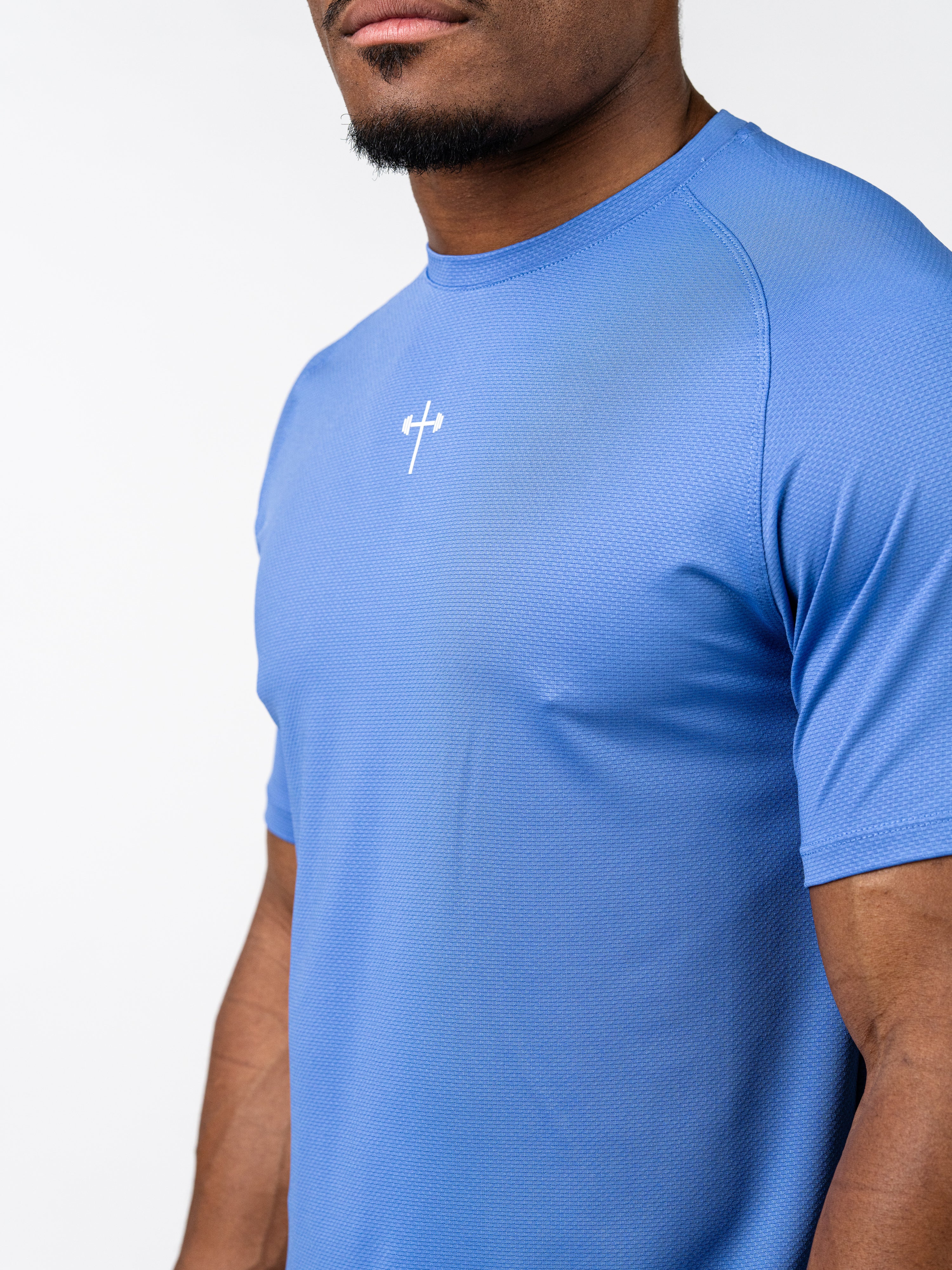 Man wearing blue Signature Tee with small white cross and barbell logo on chest, made from breathable, stretch performance fabric designed for intense training.