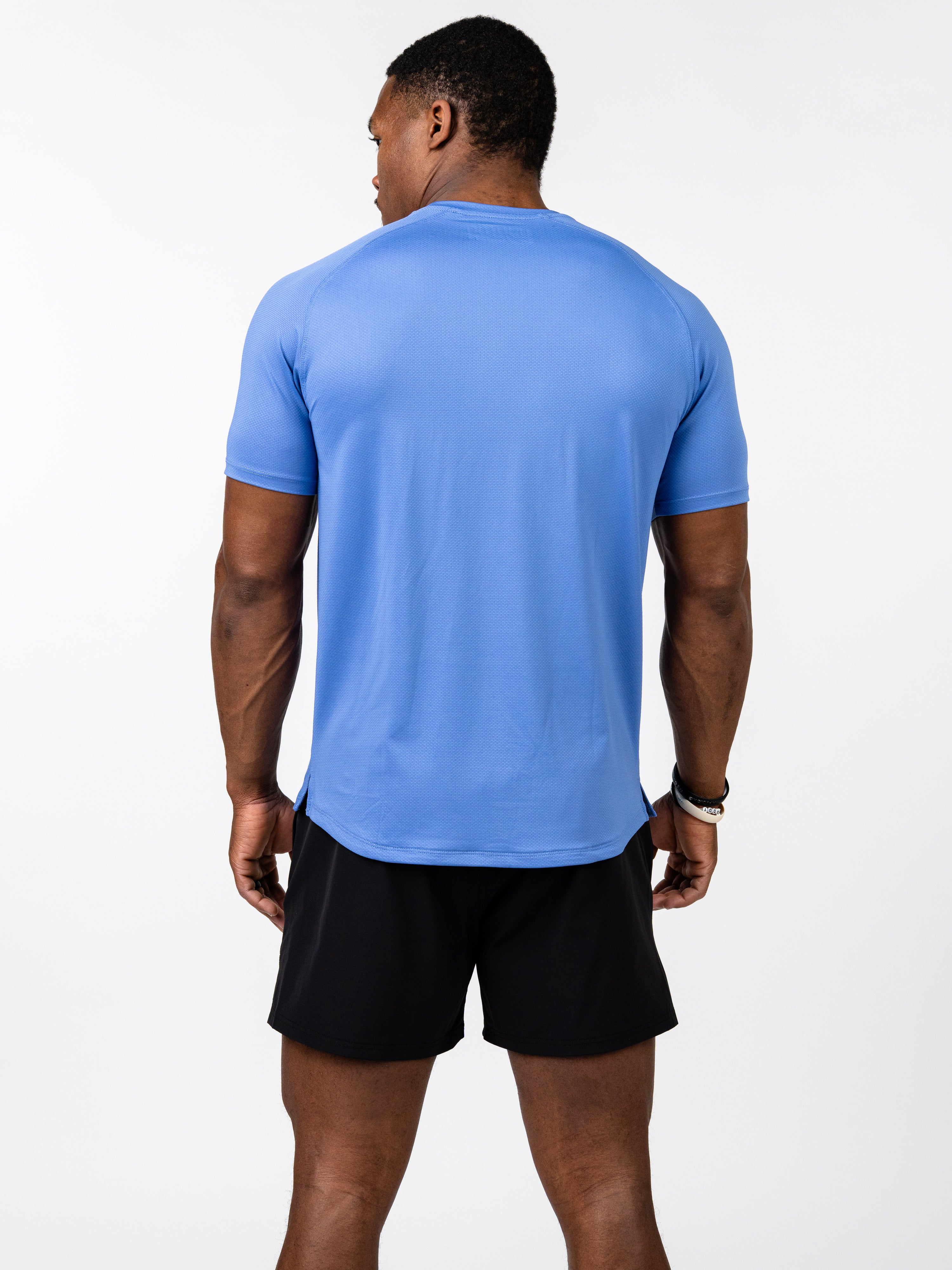 Athletic man wearing a blue Signature Tee made from breathable, stretch performance fabric, paired with black shorts, standing with back facing the camera.