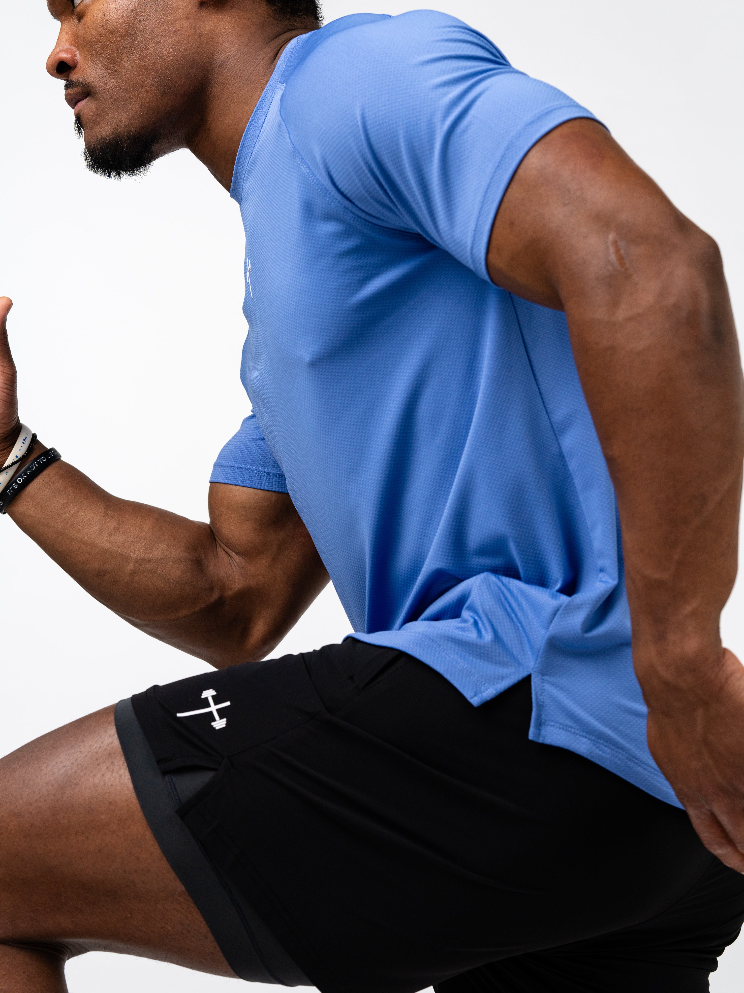 Male athlete wearing a blue Signature Tee and black shorts featuring a barbell cross symbol, demonstrating dynamic motion in a fitness setting.