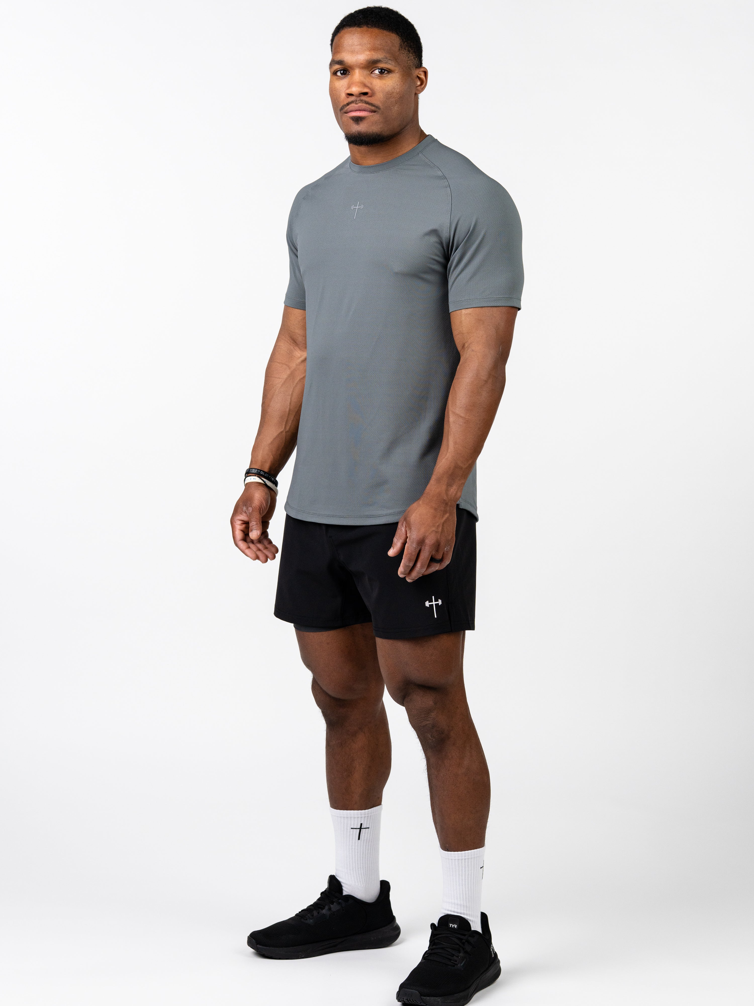Man wearing a grey Signature Tee made with breathable, sweat-wicking fabric paired with black shorts featuring a subtle cross and barbell logo, styled for Christian gym clothing.