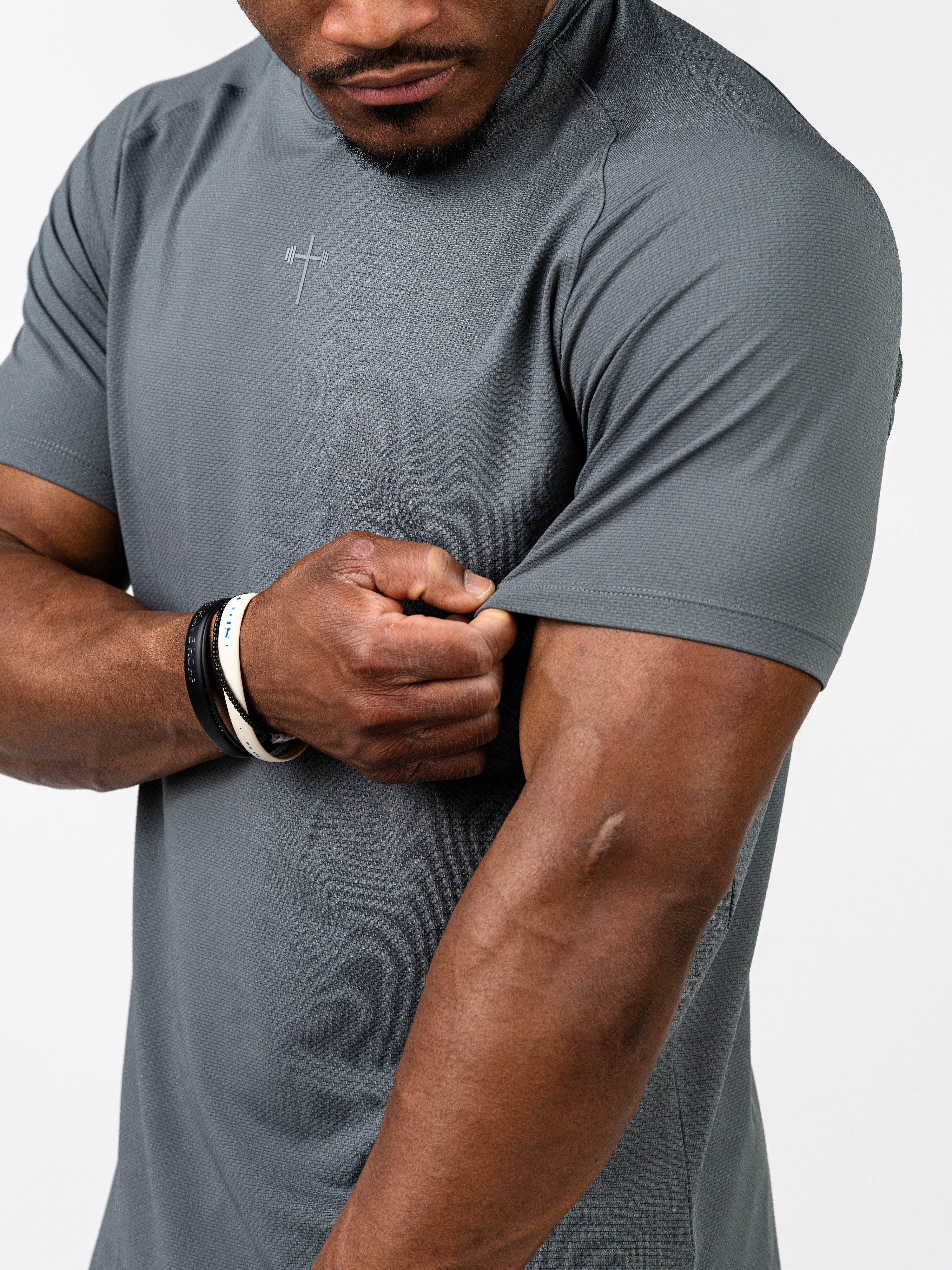 Man wearing grey Signature Tee with subtle cross and barbell design on chest, showcasing breathable, stretchy fabric and muscular arm in fitness pose.