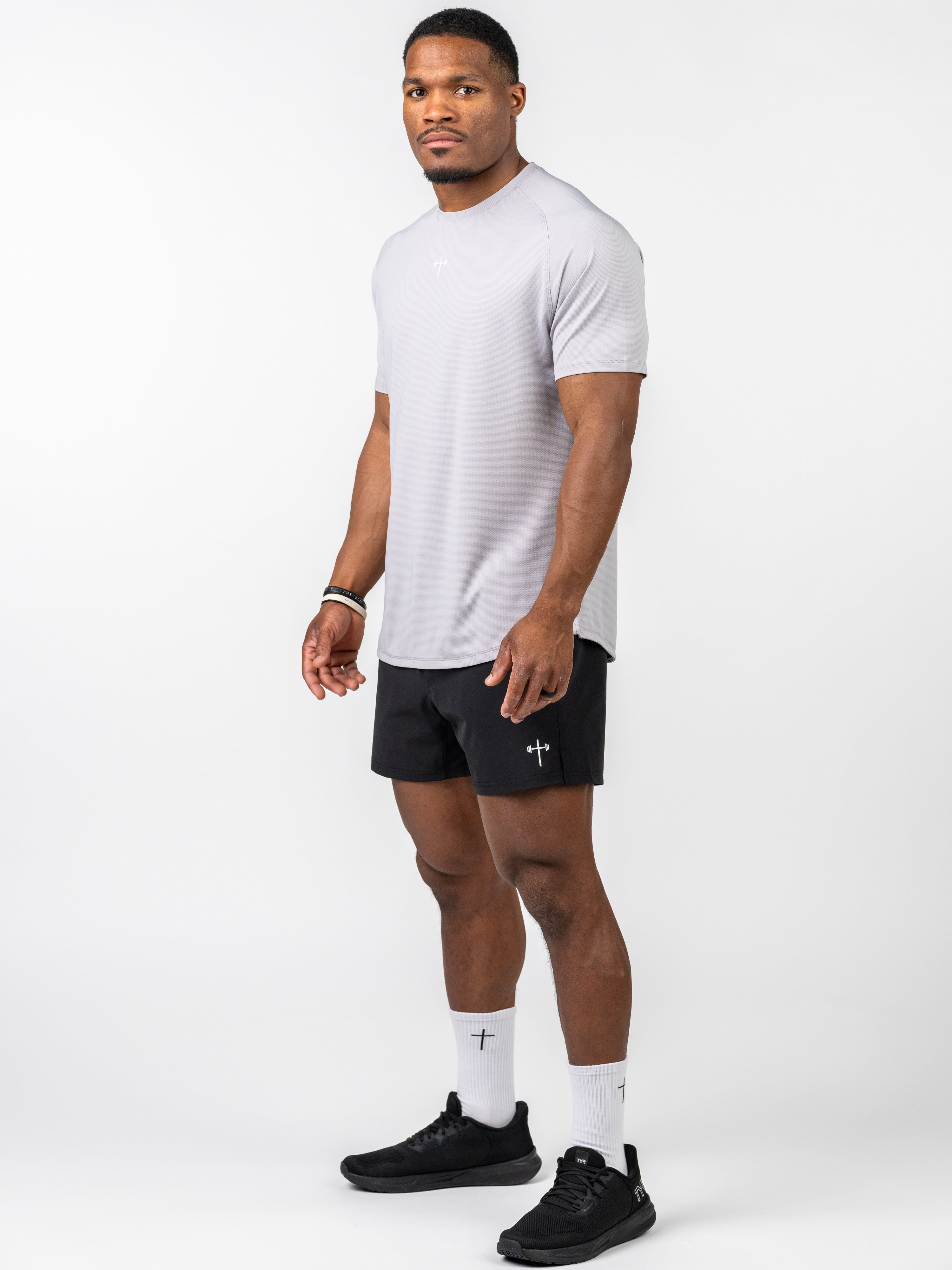 Athletic man wearing a light grey Signature Tee paired with black shorts featuring a subtle cross and barbell design, styled for Christian fitness apparel.