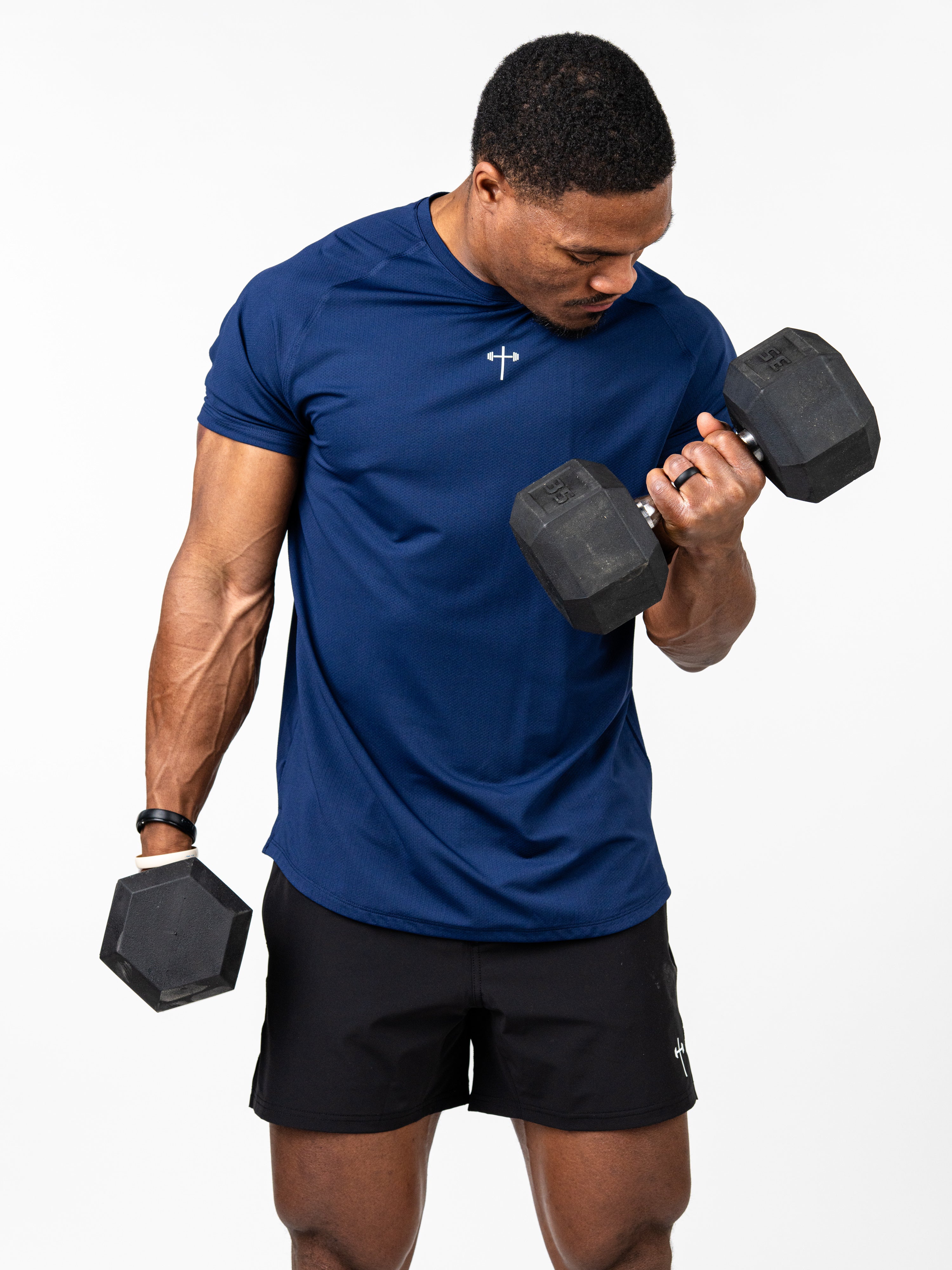 Athletic man lifting dumbbells wearing a navy blue Signature Tee with a subtle Christian cross and barbell design, paired with black workout shorts.