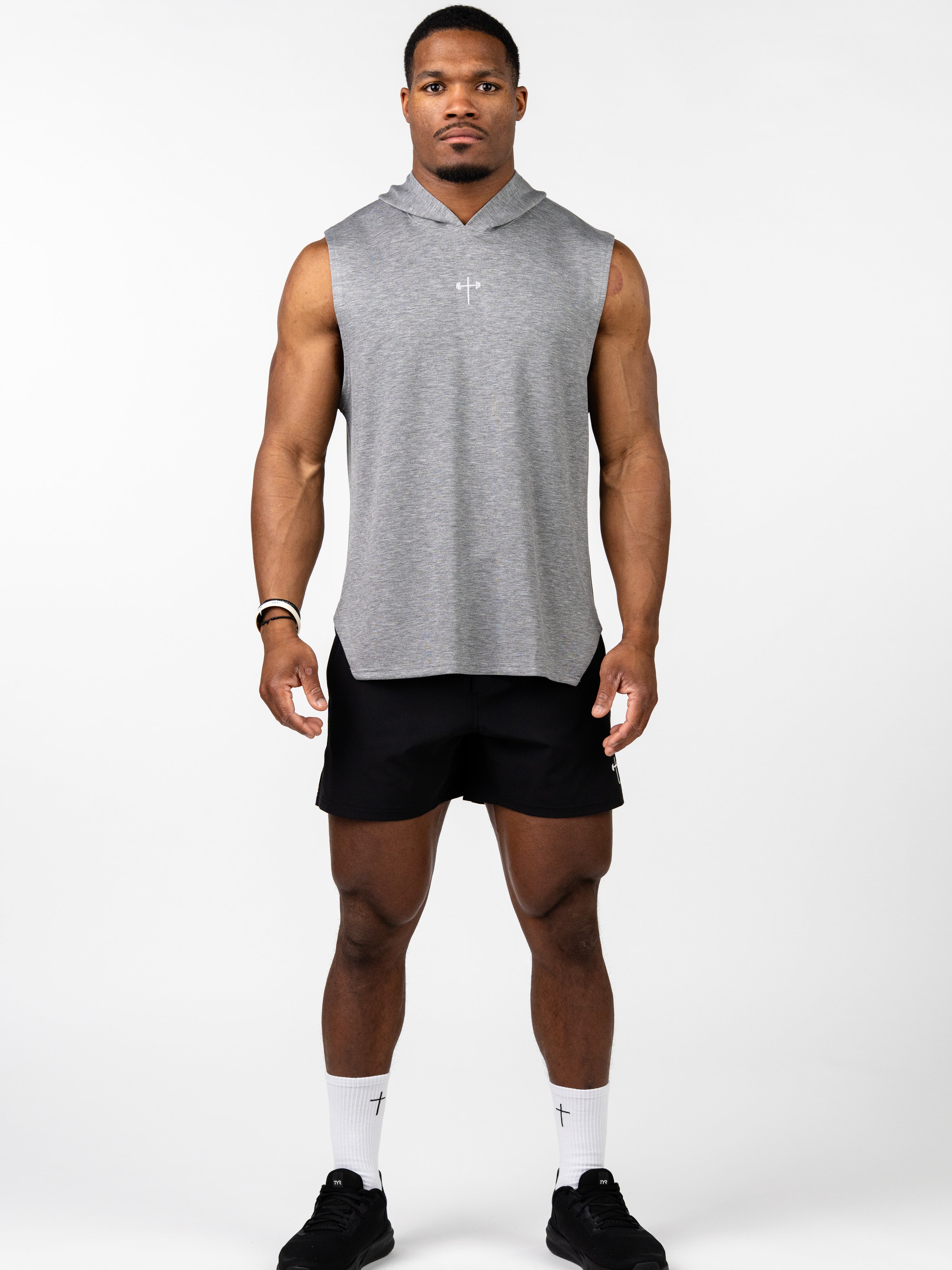 Athletic man wearing a gray Essential Sleeveless Hoodie with a small white cross logo on the chest, paired with black shorts, white socks featuring a cross, and black sneakers.