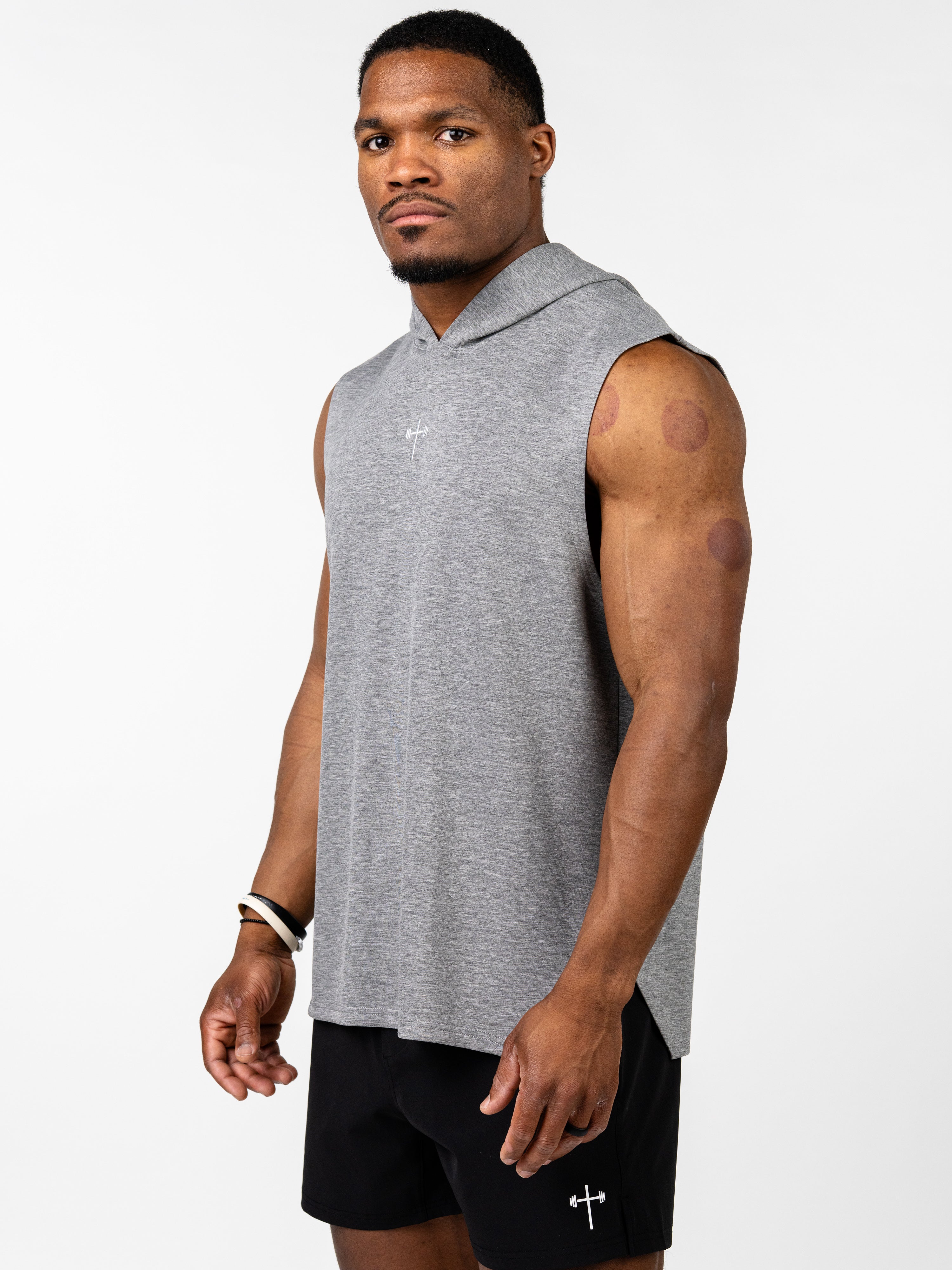 Athletic man wearing a gray Essential Sleeveless Hoodie with a subtle Christian cross design, paired with black shorts featuring a small barbell cross logo.