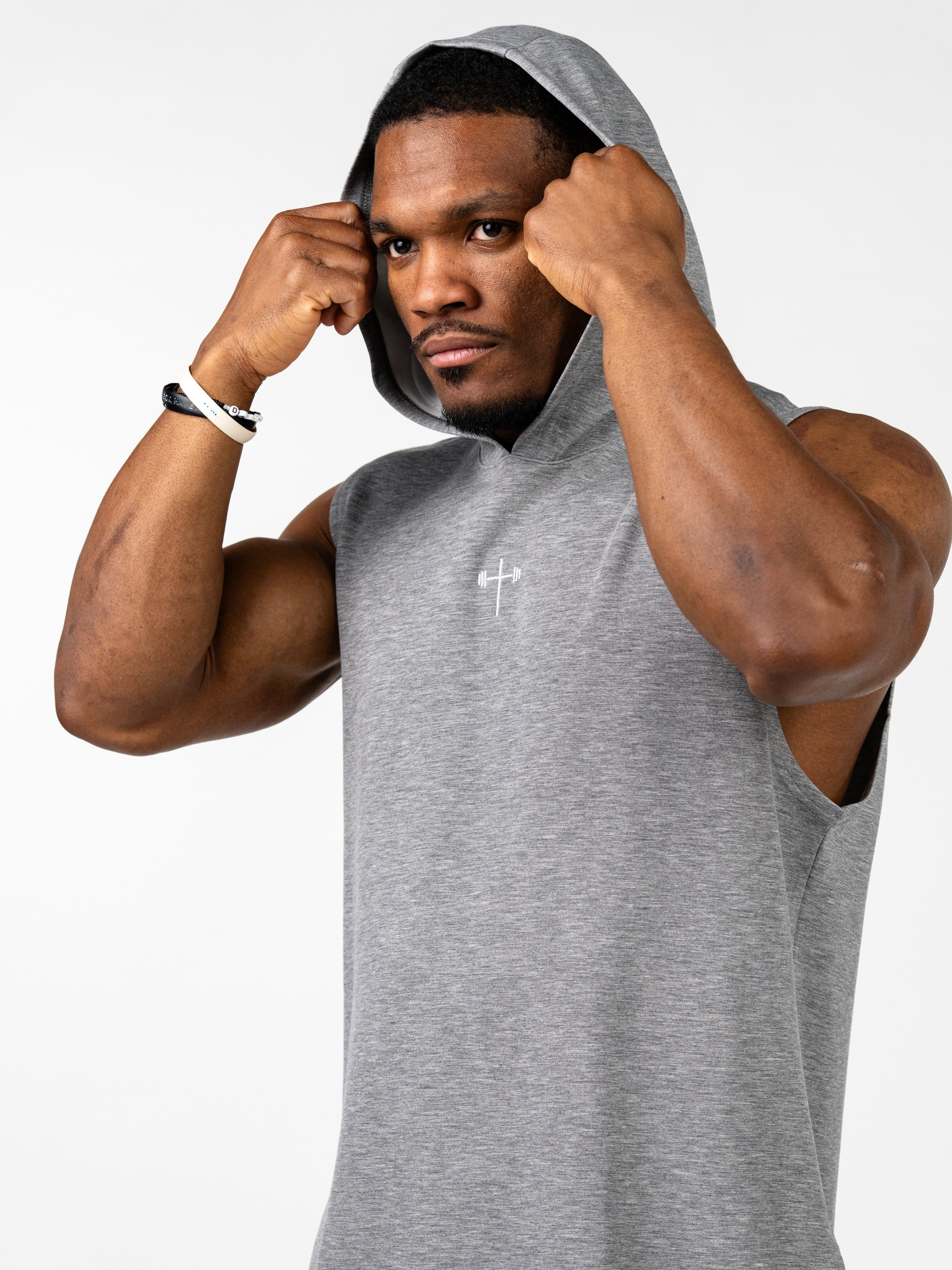 Athletic man wearing a gray Essential Sleeveless Hoodie with a small white cross and barbell design on the chest, adjusting the hood in a studio setting.