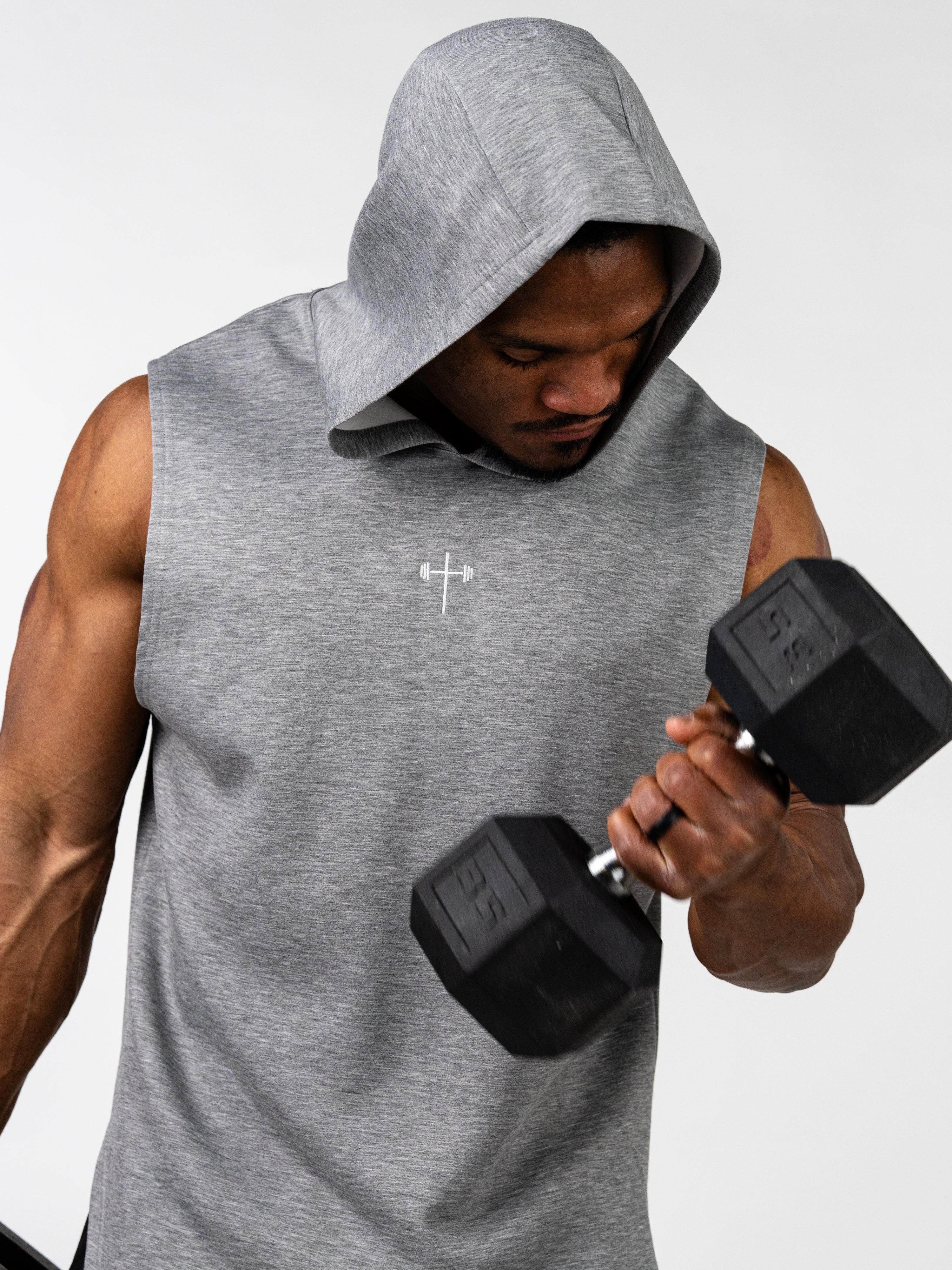 Athletic man wearing a gray Essential Sleeveless Hoodie with a small Christian cross and barbell logo, lifting a dumbbell during a workout session.