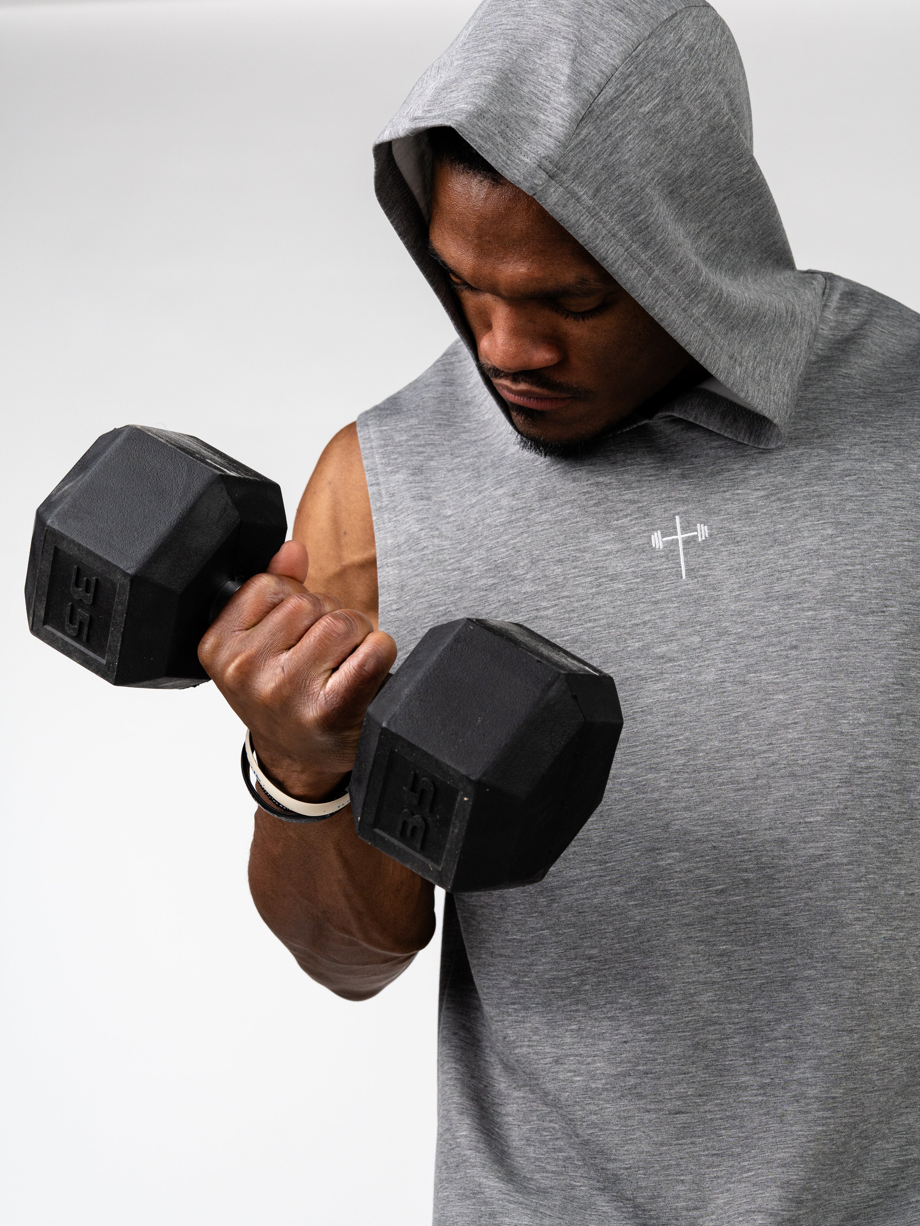 Athlete wearing a grey Essential Sleeveless Hoodie with a small white barbell cross logo, lifting a 35-pound dumbbell while looking down, demonstrating strength and focus.