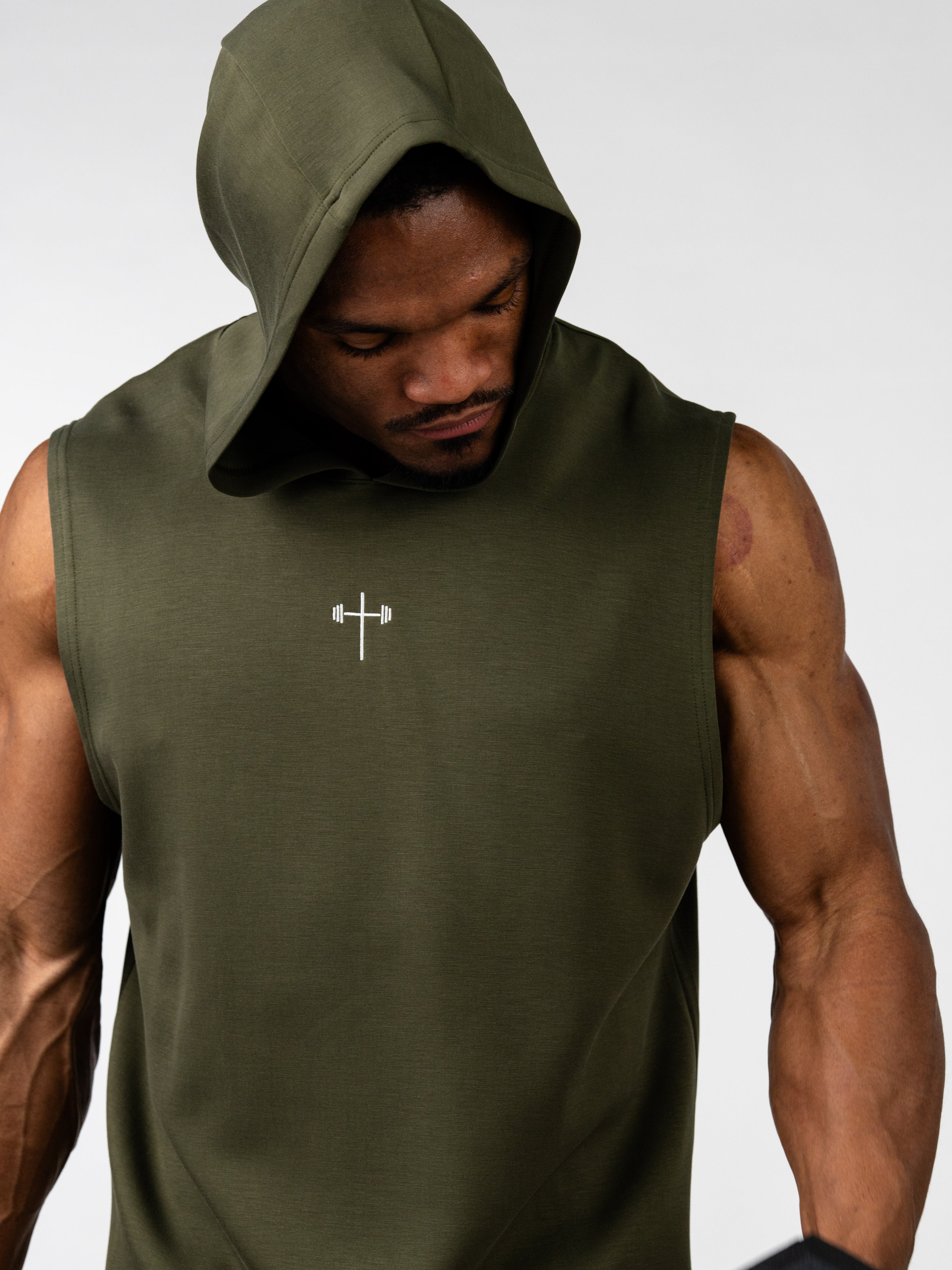 Man wearing an olive green Essential Sleeveless Hoodie with a hood up, featuring a small white Christian cross and barbell logo on the chest, showcasing muscular arms.