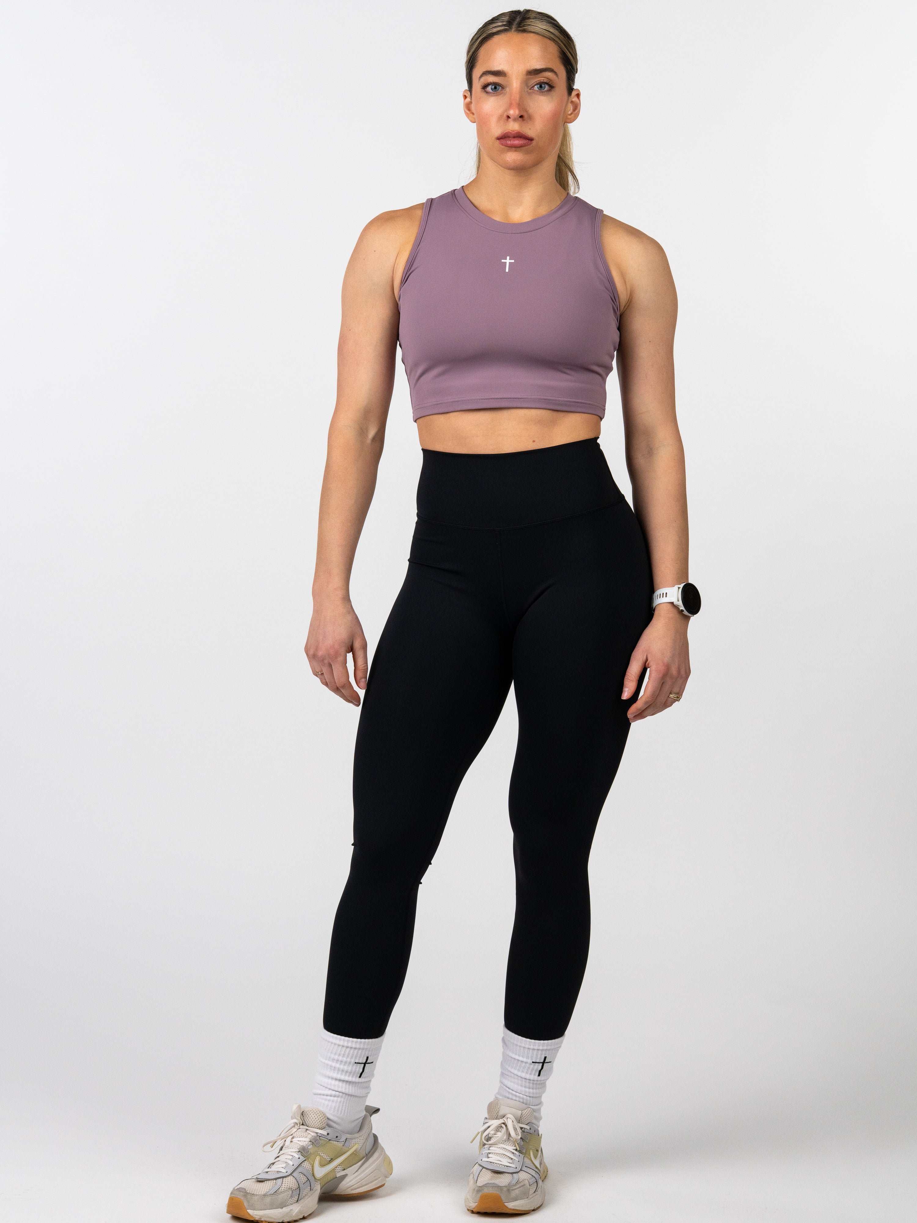 Woman wearing a mauve Vital Cross Combat Top with a small white cross on the chest, paired with black leggings, white socks with cross detail, and athletic shoes.