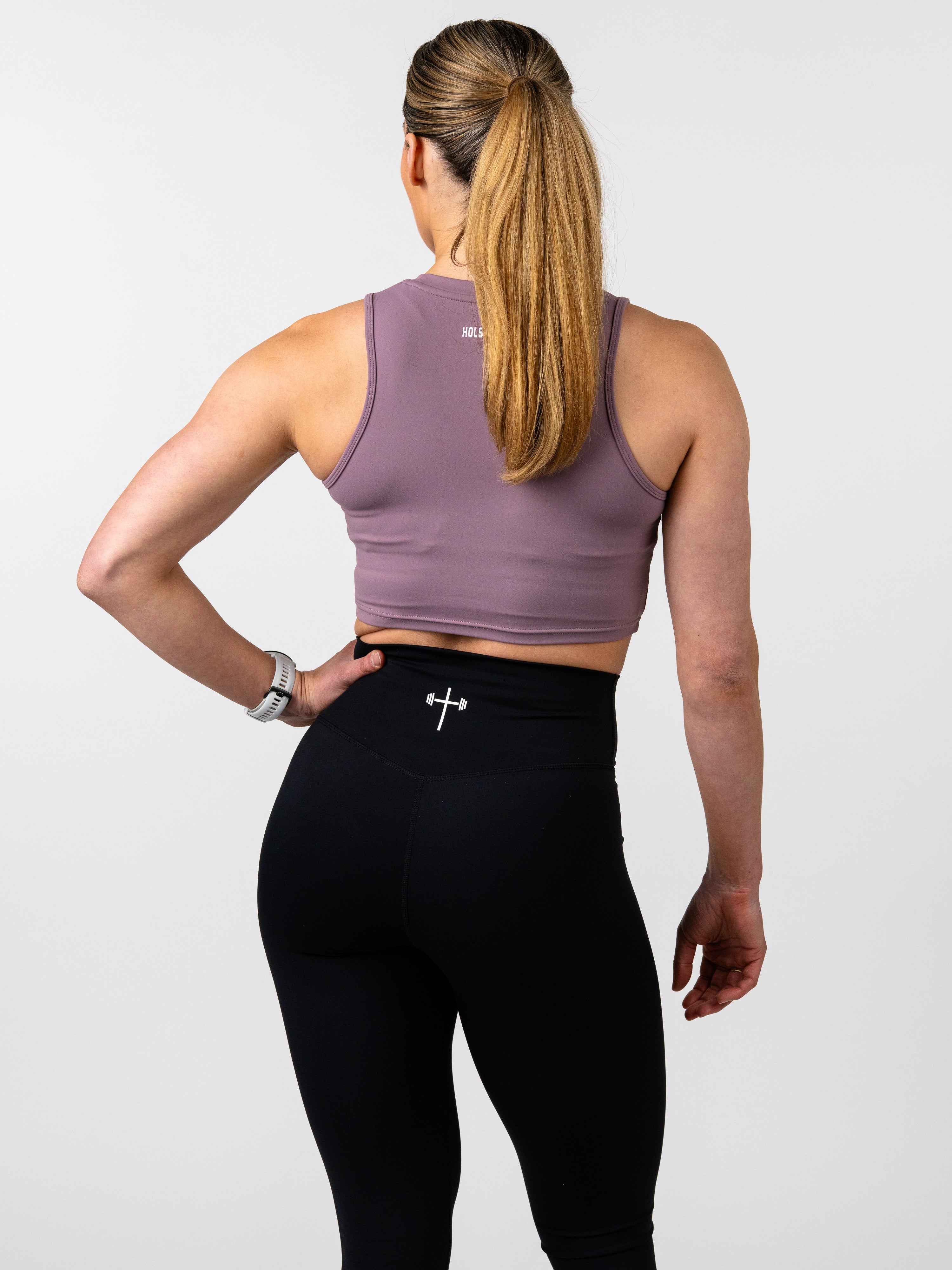 Woman seen from the back wearing the Vital Cross Combat Top in mauve paired with black leggings, showcasing the fitted, athletic design and subtle Christian cross with barbell logo.