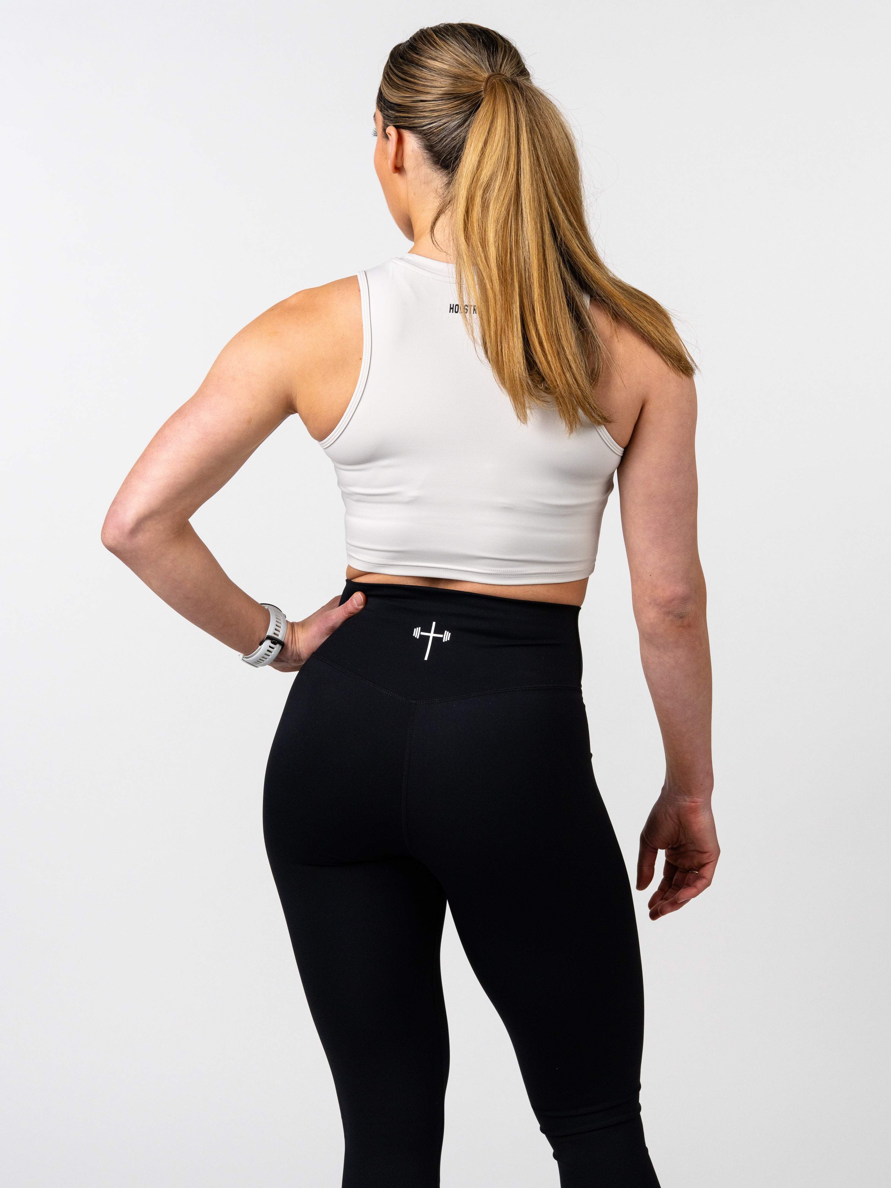 Woman wearing a white Vital Cross Combat Top and black leggings, shown from behind, highlighting the top’s fitted design and Christian cross with barbell detail on the waistband.