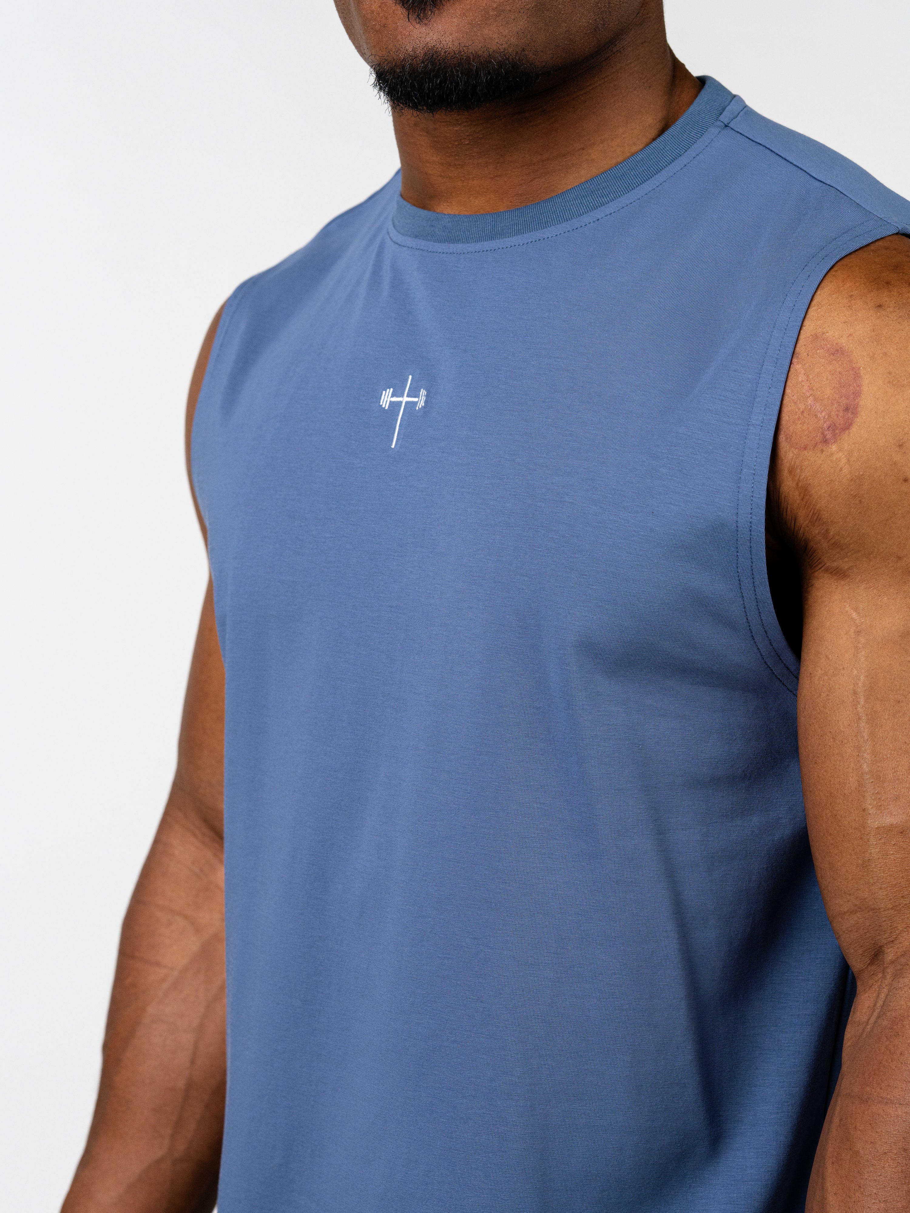 Close-up of a man wearing a blue Vital Cross Sleeveless Tank featuring a small white cross with a barbell design at the center chest, designed for Christian gym clothing and activewear.