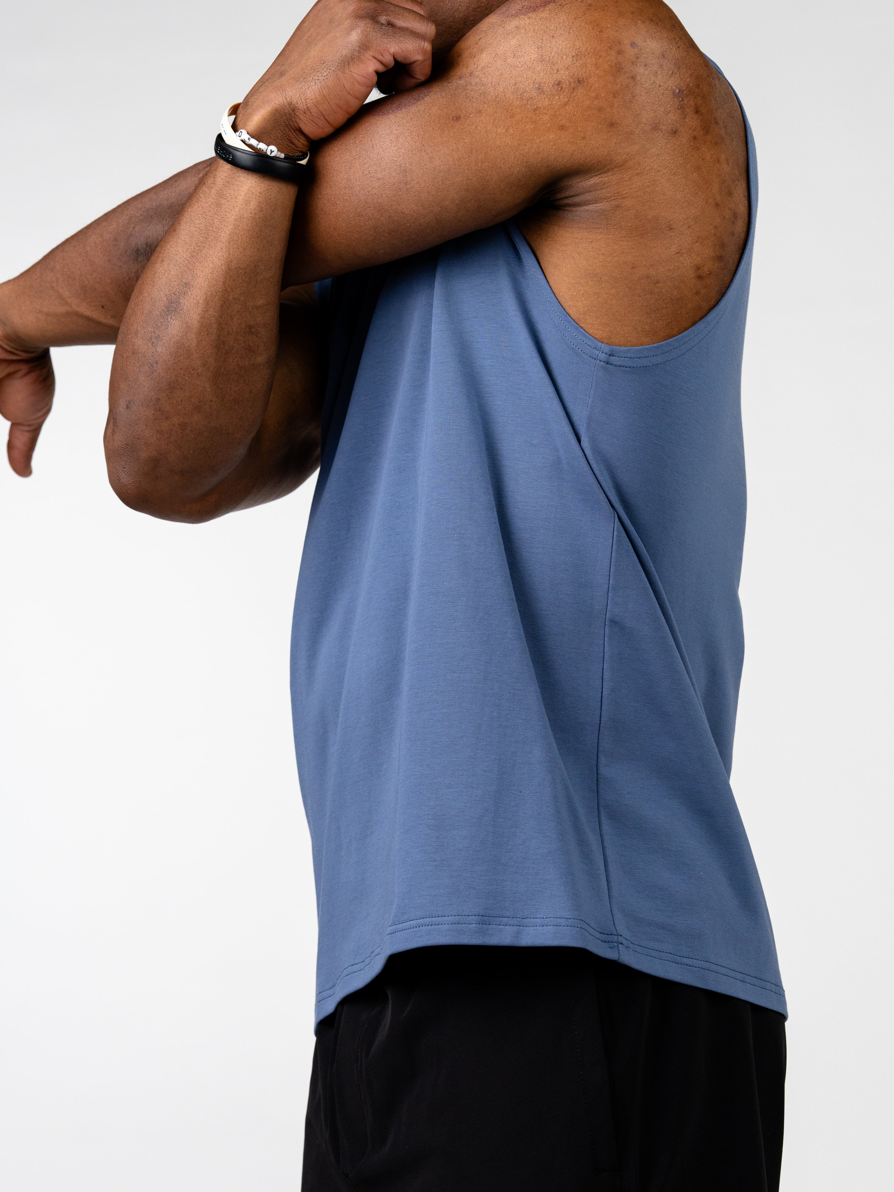 Man stretching arm wearing a blue Vital Cross Sleeveless Tank designed for high-output training and unrestricted movement, paired with black shorts against a white background.