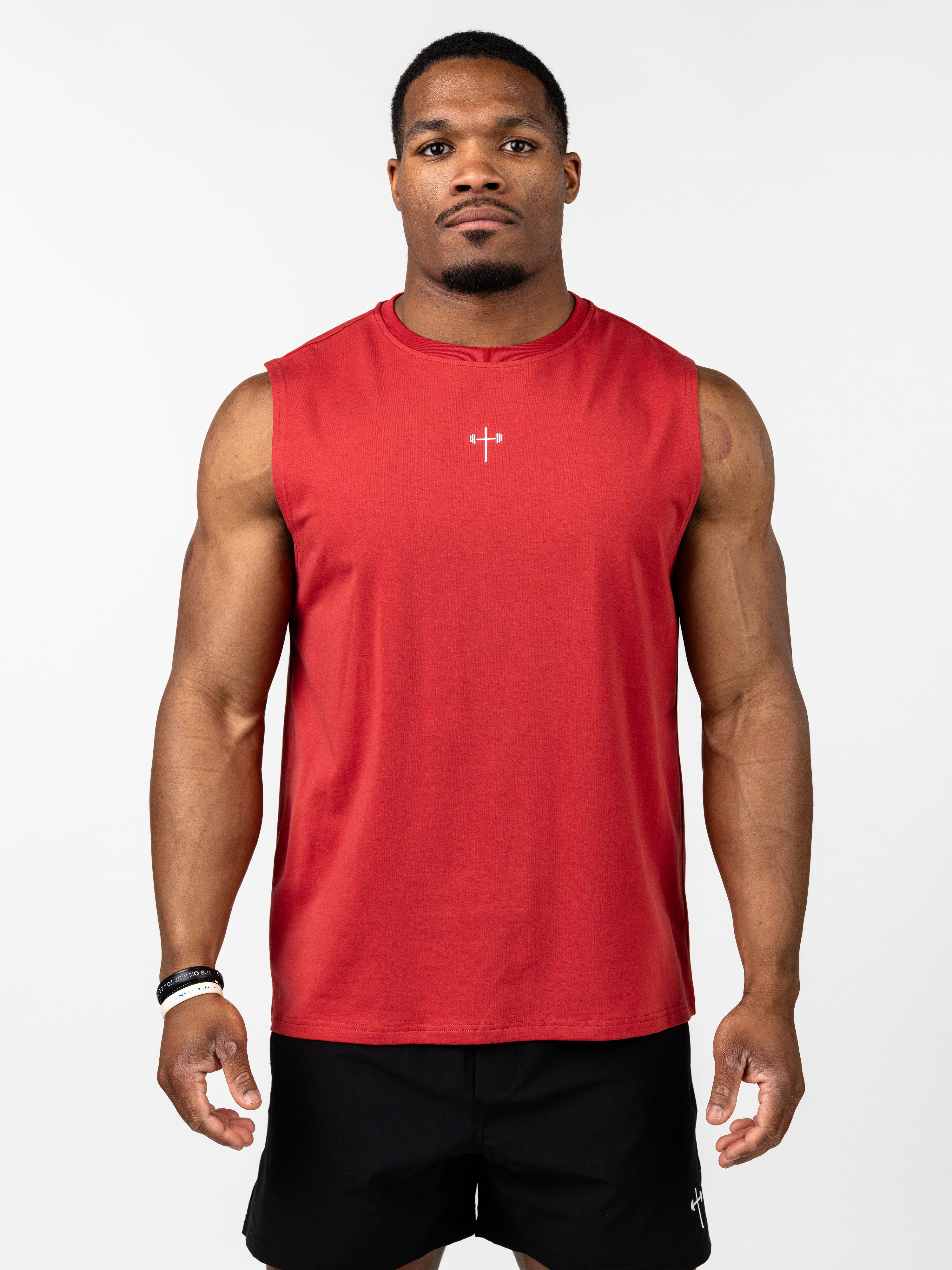 Male model wearing a red Vital Cross Sleeveless Tank designed for high-output training, featuring a small white Christian cross with a barbell emblem on the chest, paired with black shorts.