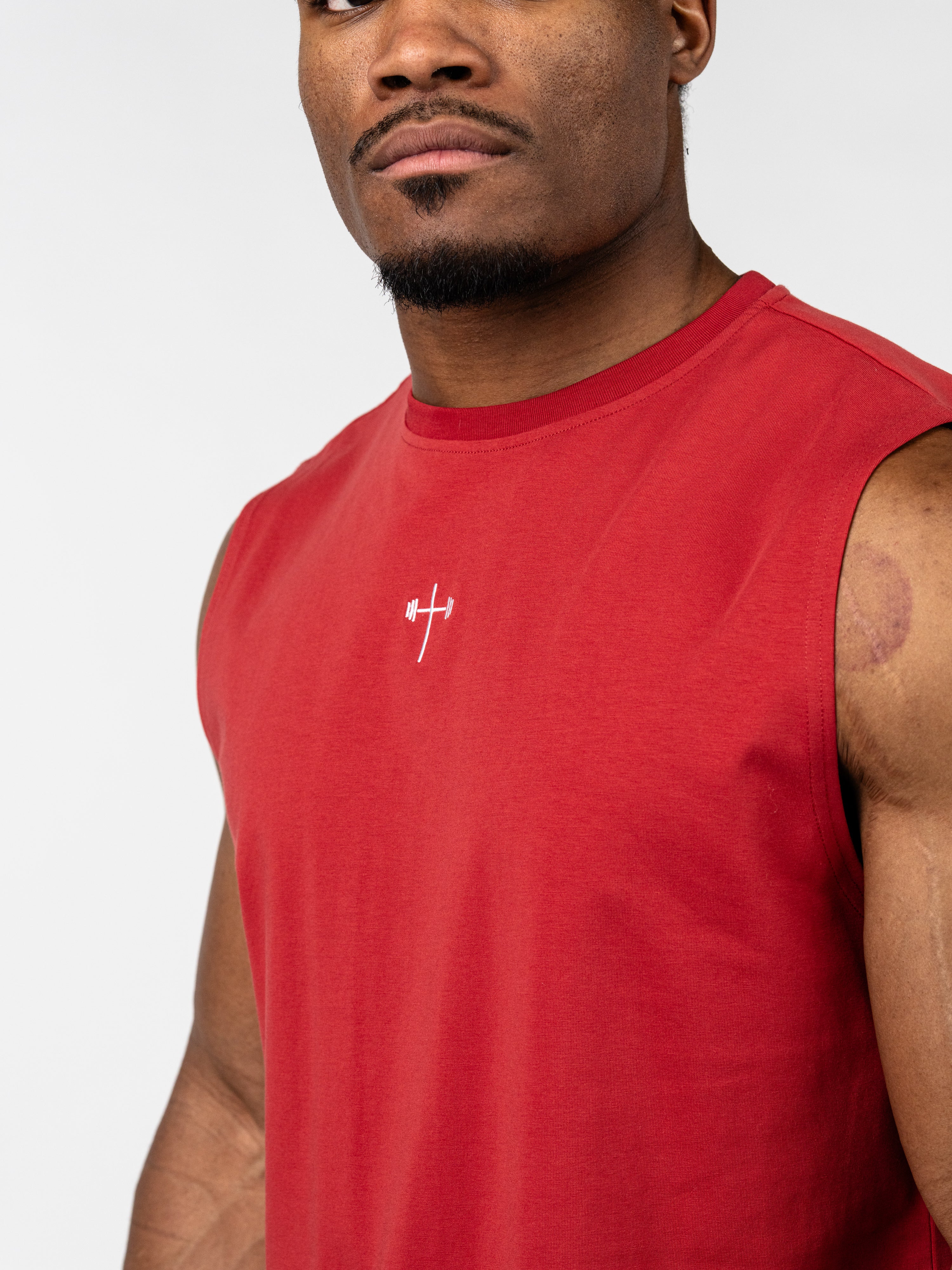 Man wearing a red Vital Cross Sleeveless Tank featuring a small white cross with a barbell graphic on the chest, representing Christian fitness apparel for high-intensity workouts.