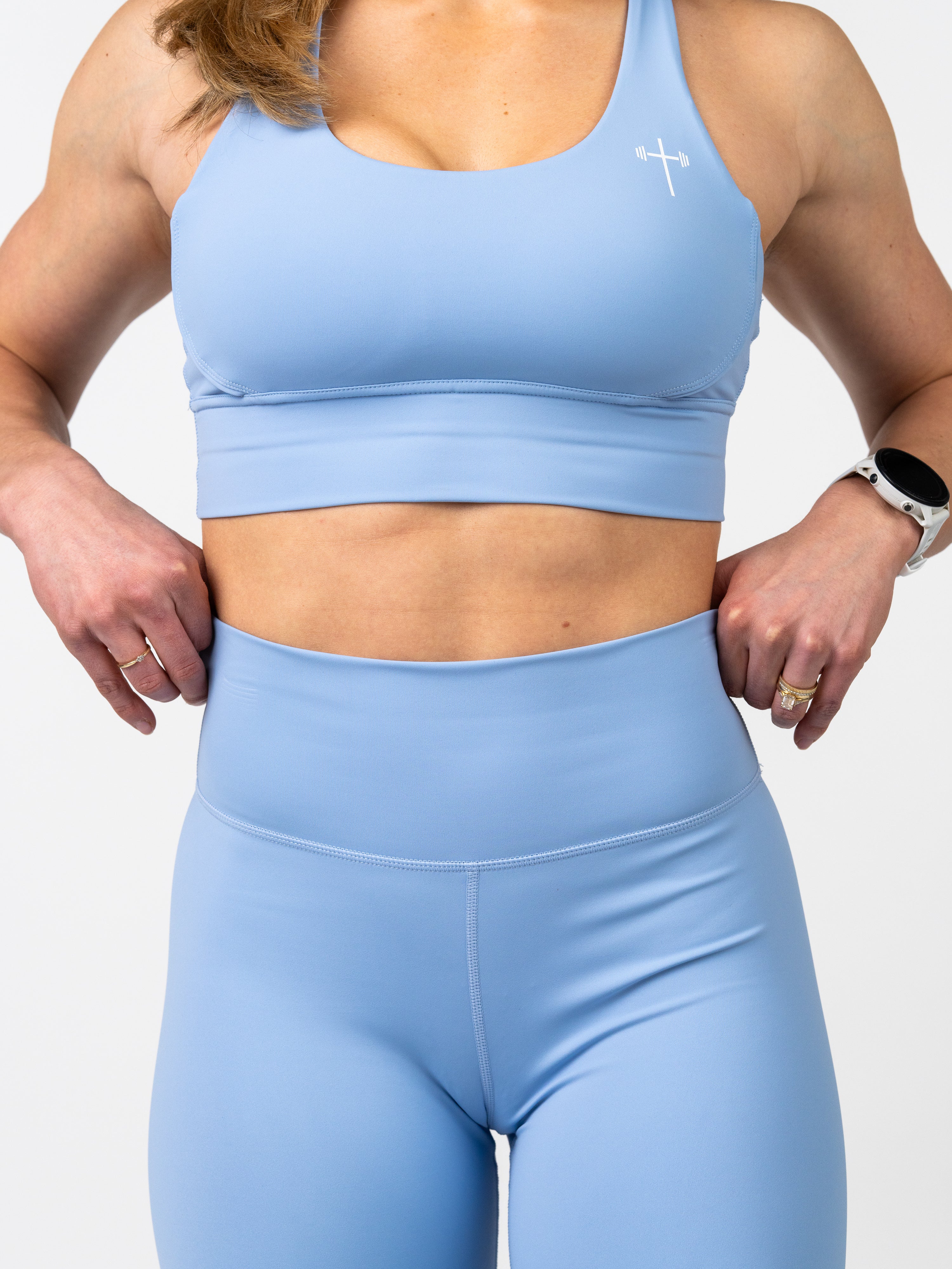 Woman wearing a light blue Essential Sports Bra with a small white cross and barbell logo on the chest, paired with matching high-waisted leggings, showing fit and comfort.