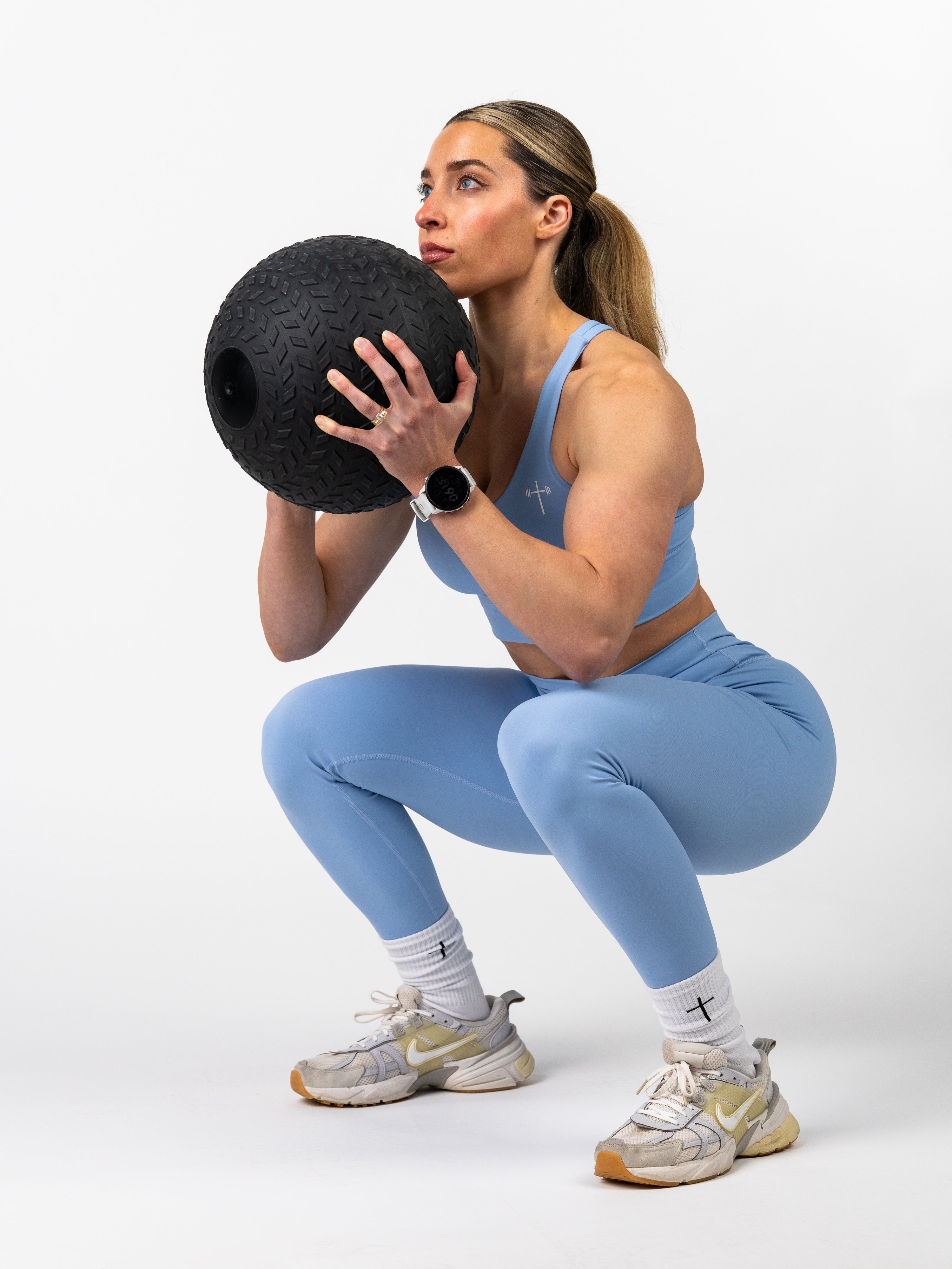 Woman wearing an Essential Sports Bra in light blue, performing a squat exercise holding a textured black medicine ball, showcasing Christian fitness apparel with cross logo details.