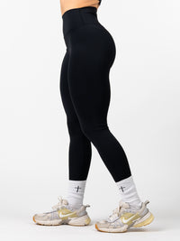 Side view of a person wearing black Essential Leggings made from a premium nylon/spandex blend, paired with white socks featuring black crosses and beige athletic shoes.