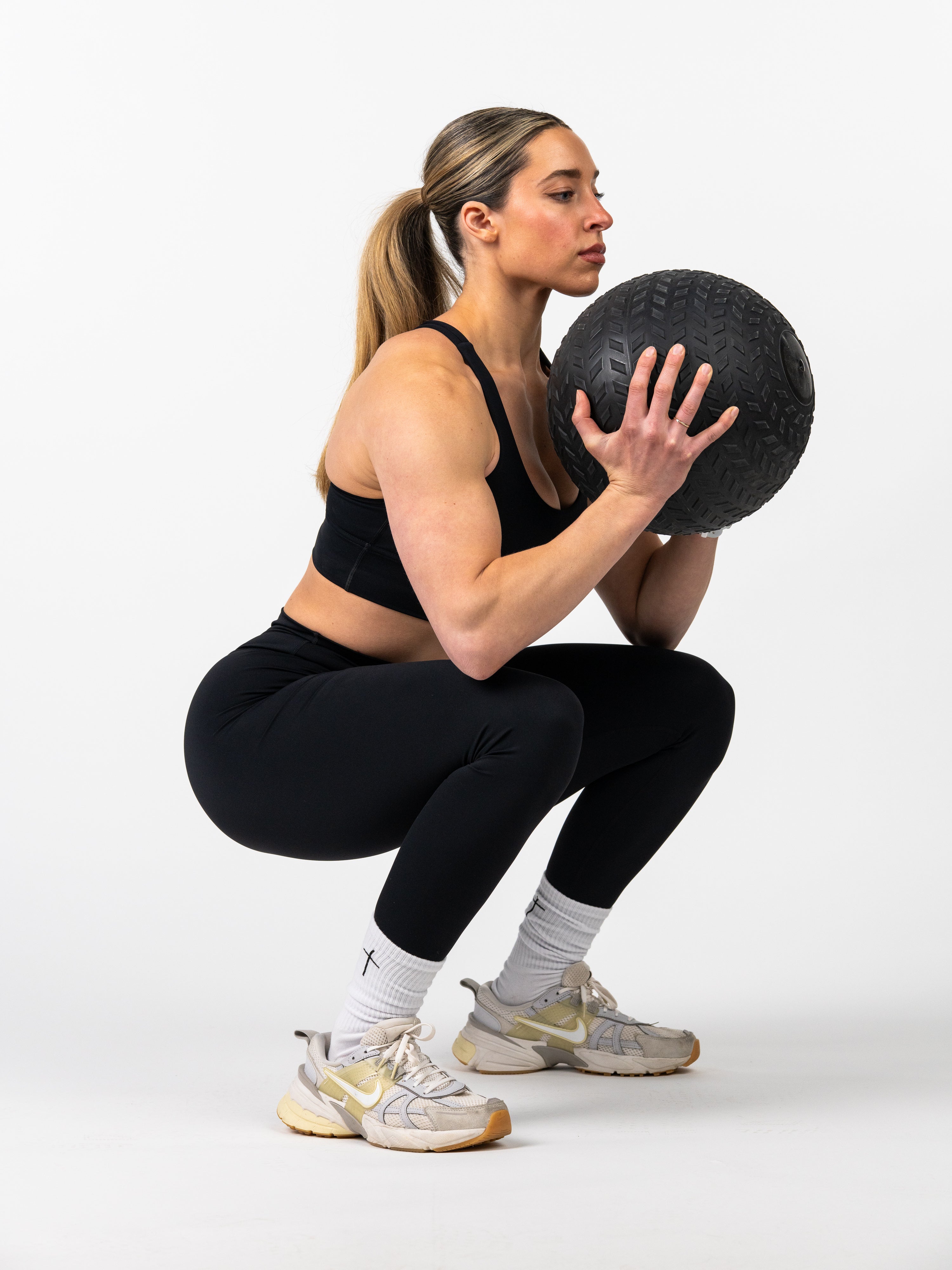 Athletic woman wearing Essential Leggings and a sports bra performing a weighted squat exercise holding a textured black medicine ball against a white background.