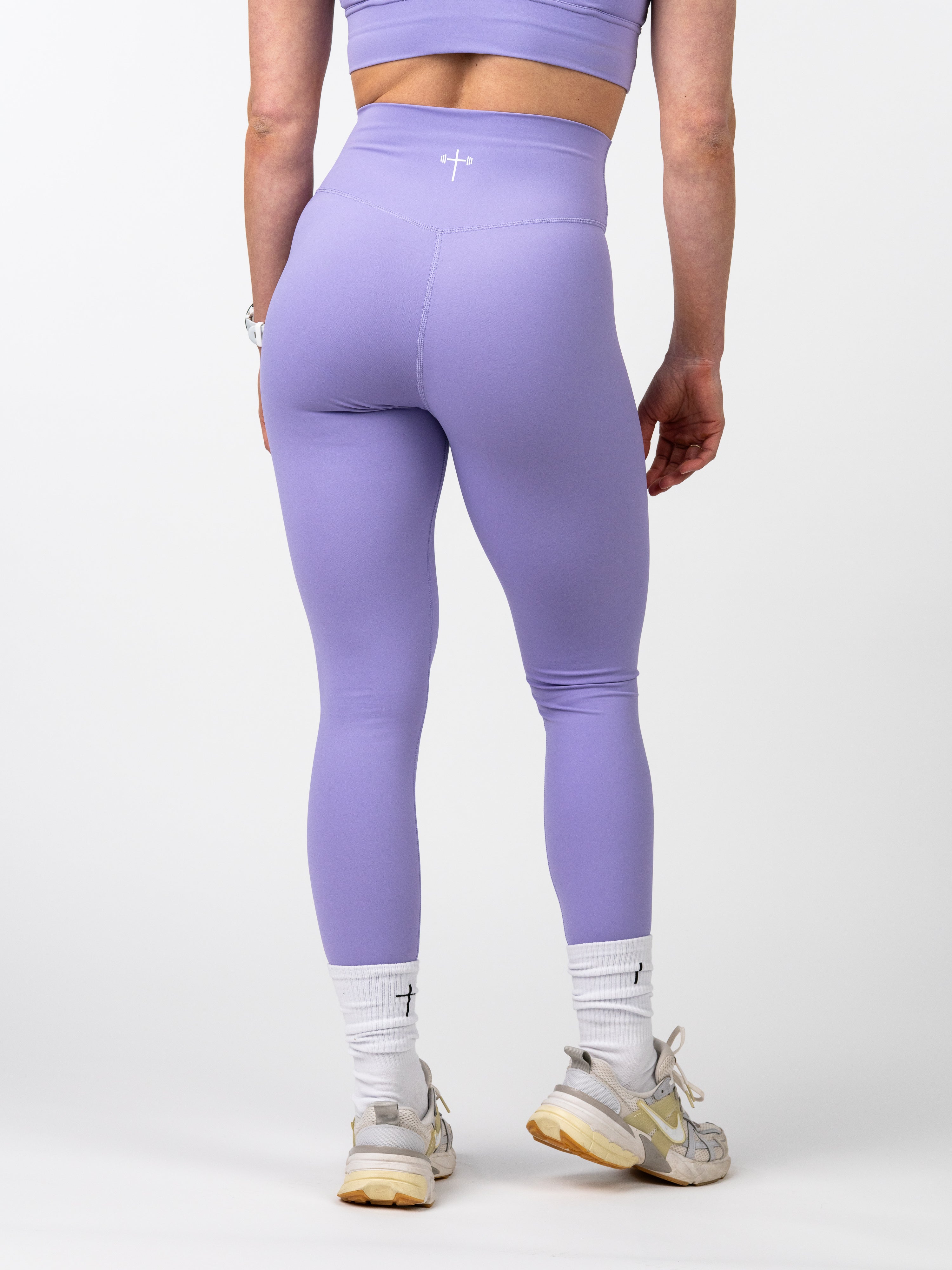Rear view of a person wearing lavender Essential Leggings, featuring a high waistband and a small Christian cross logo, paired with white socks and sneakers.