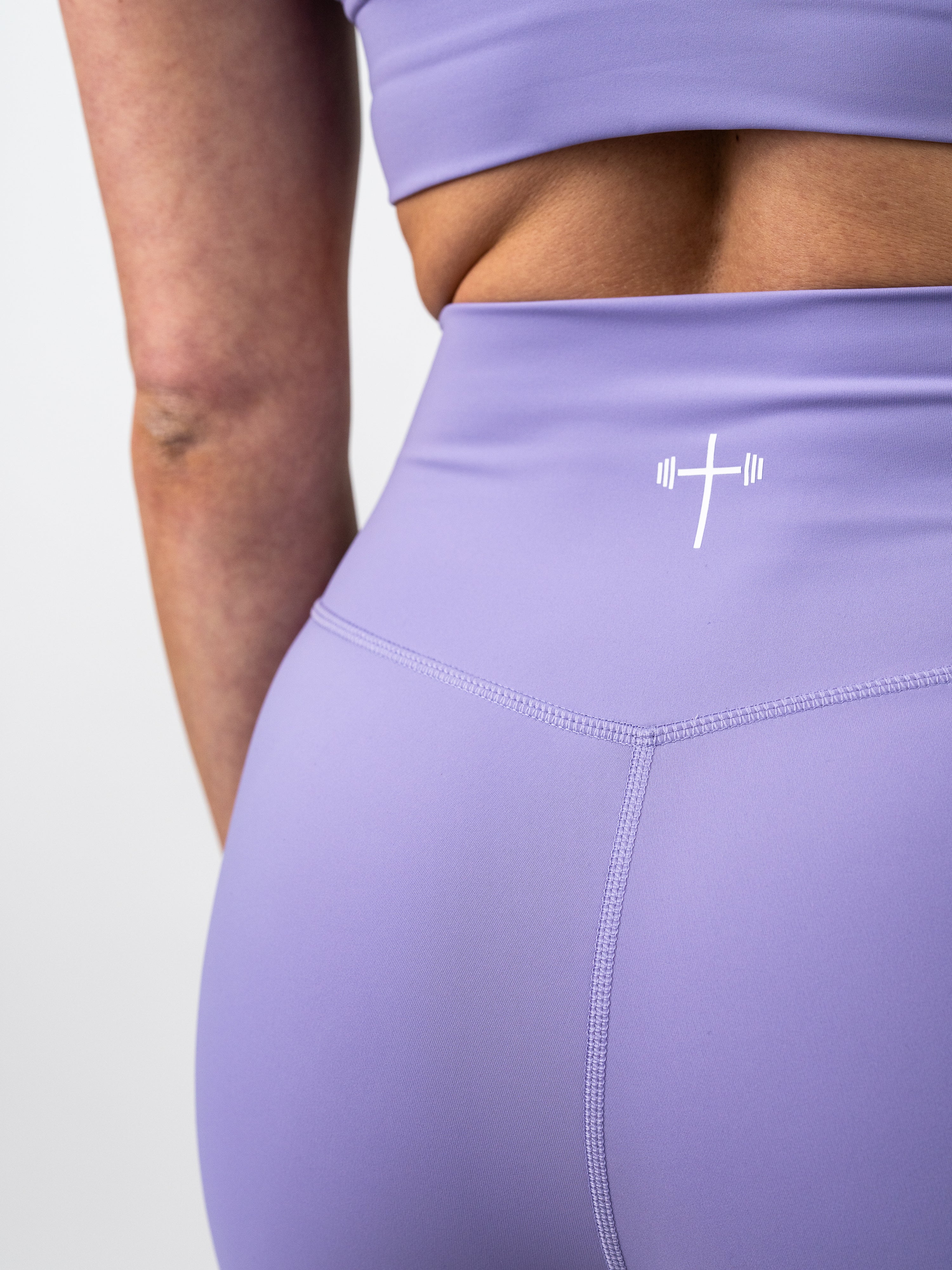 Close-up view of a person wearing lavender Essential Leggings featuring a small white Christian cross with a barbell logo on the high waistband, designed for endurance and flexibility.