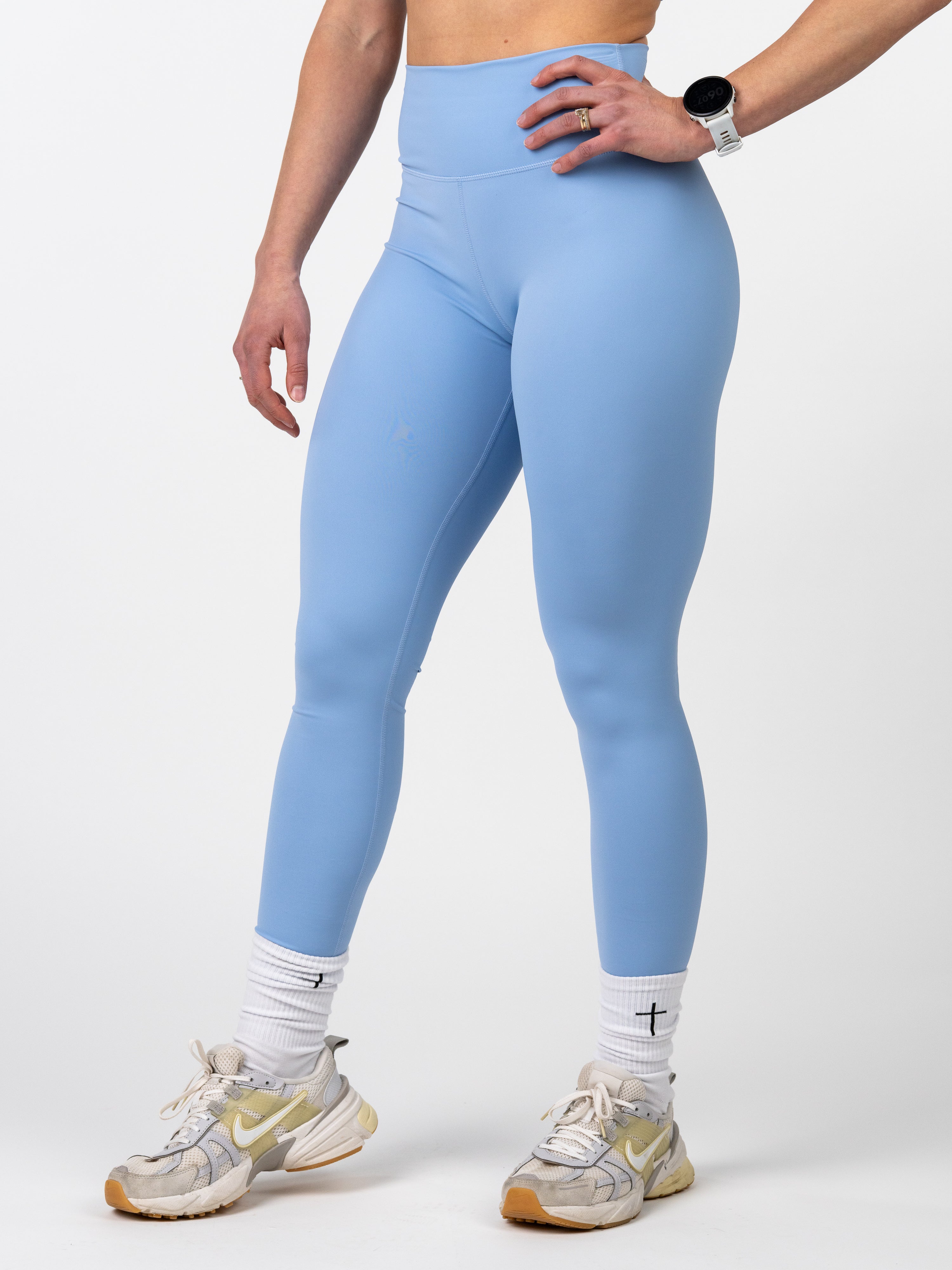 Close-up of a person wearing light blue Essential Leggings paired with white socks featuring a black cross, and white athletic shoes, standing against a plain background.