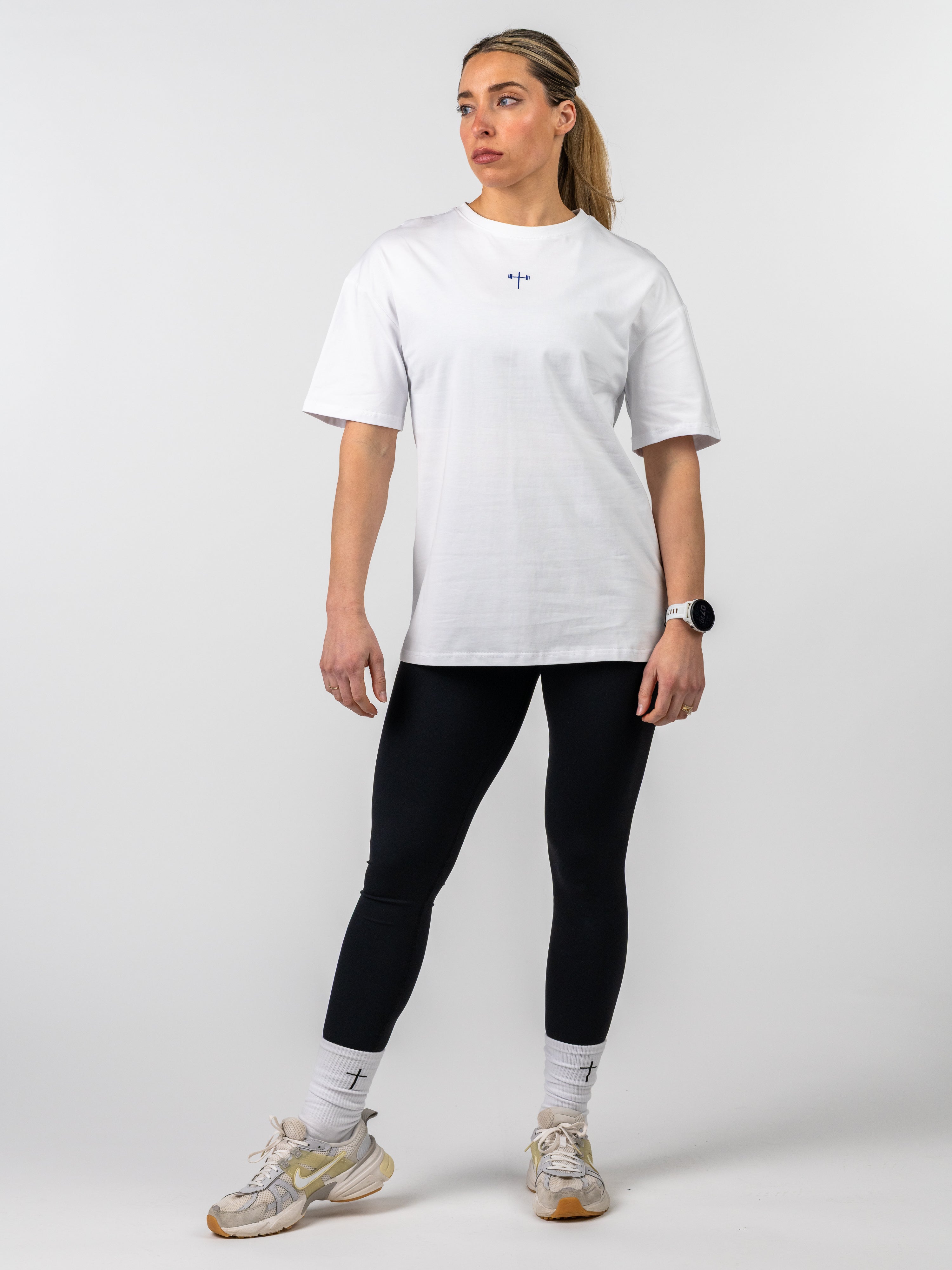 Woman modeling the Women's Lifting Club Oversized Tee in white with a small cross and barbell design, paired with black leggings and white socks with cross details.