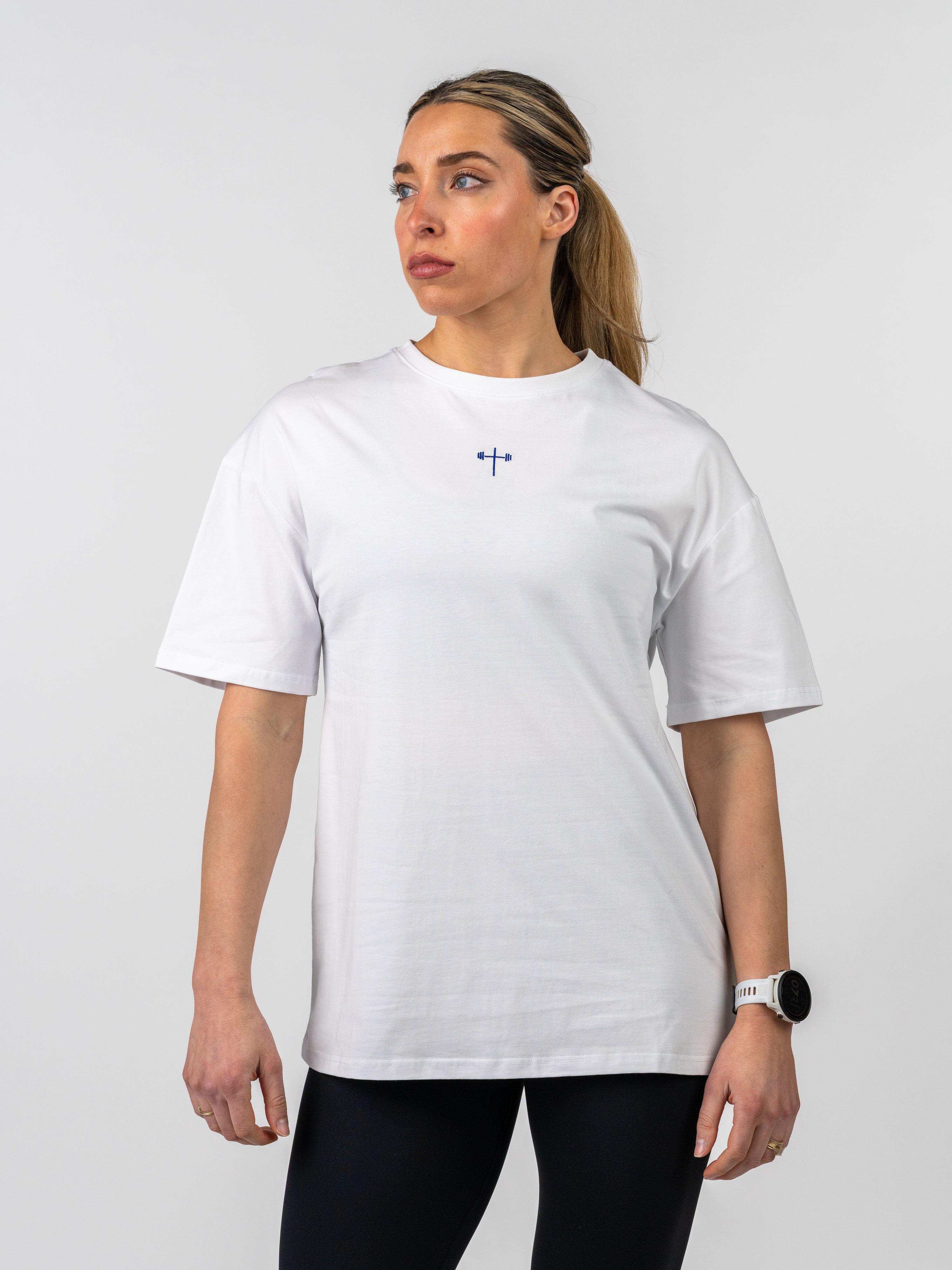 Woman wearing a white Women's Lifting Club Oversized Tee featuring a small blue cross and barbell graphic on the chest, styled with black leggings against a plain background.