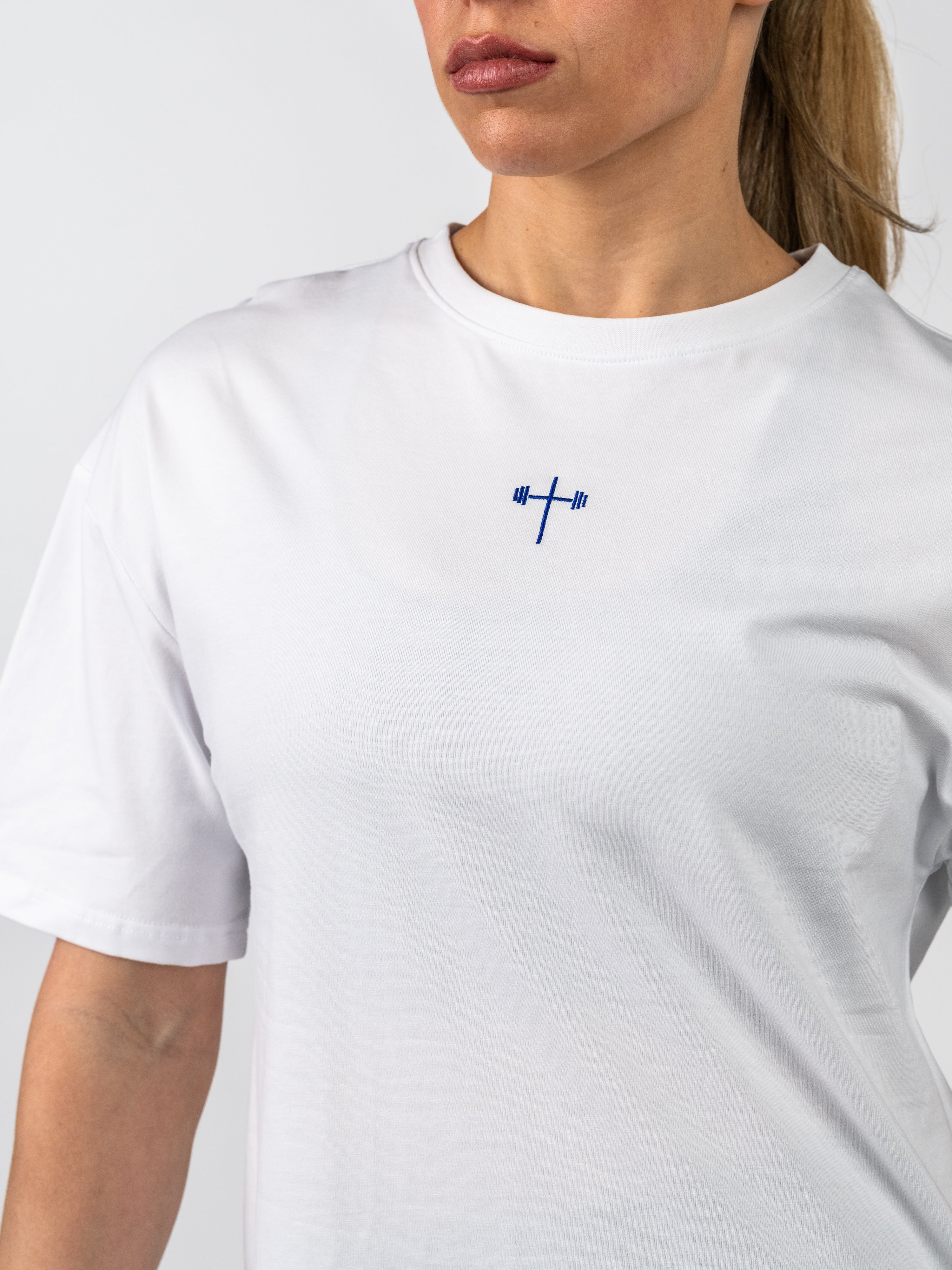 Close-up of a woman wearing a white Women's Lifting Club Oversized Tee featuring a small blue cross with a barbell design on the chest, representing Christian gym clothing with faith-based fitness inspiration.