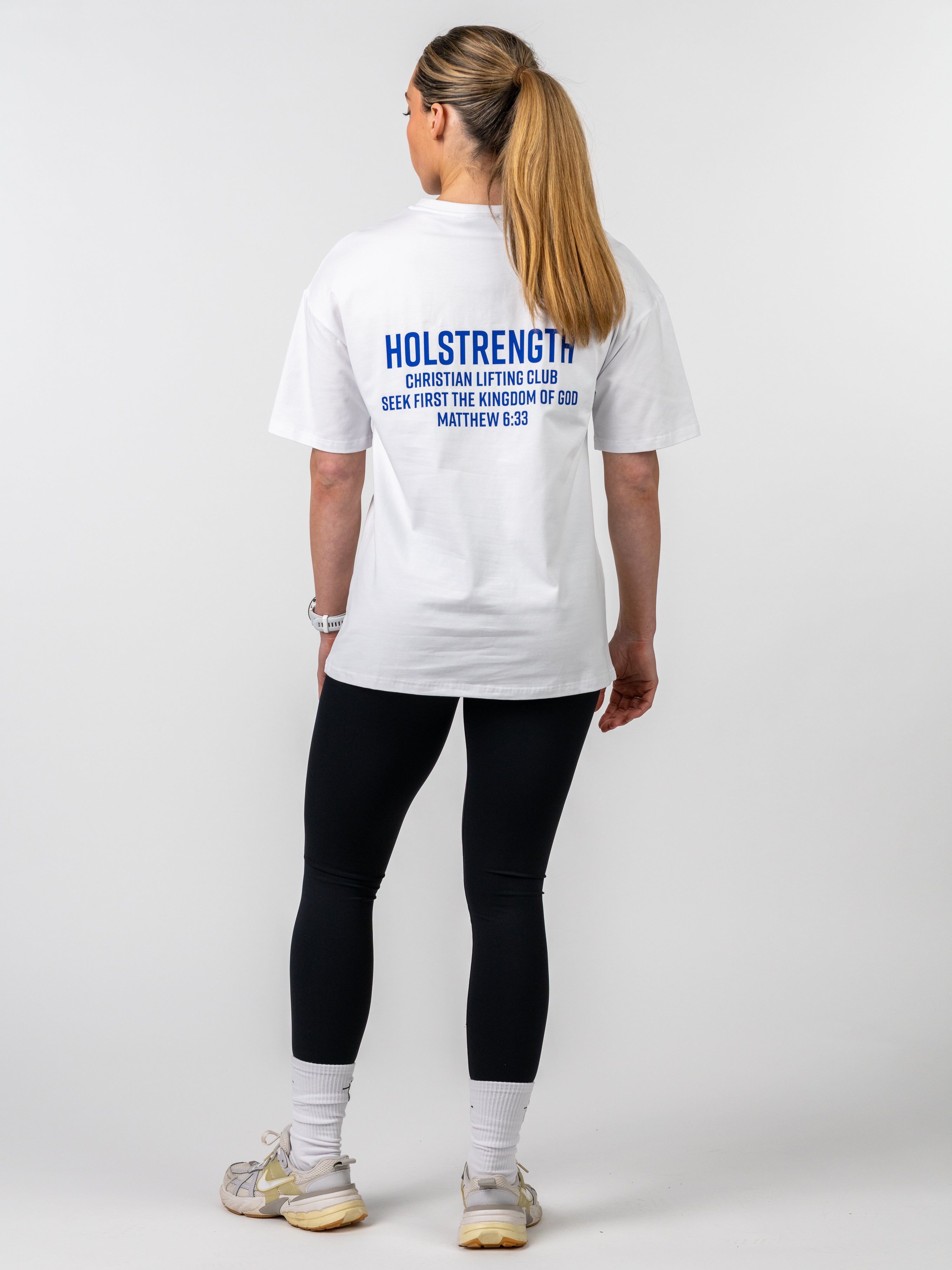 Woman wearing a white Women's Lifting Club Oversized Tee with blue text on the back promoting Christian lifting club and Matthew 6:33, paired with black leggings and sneakers.
