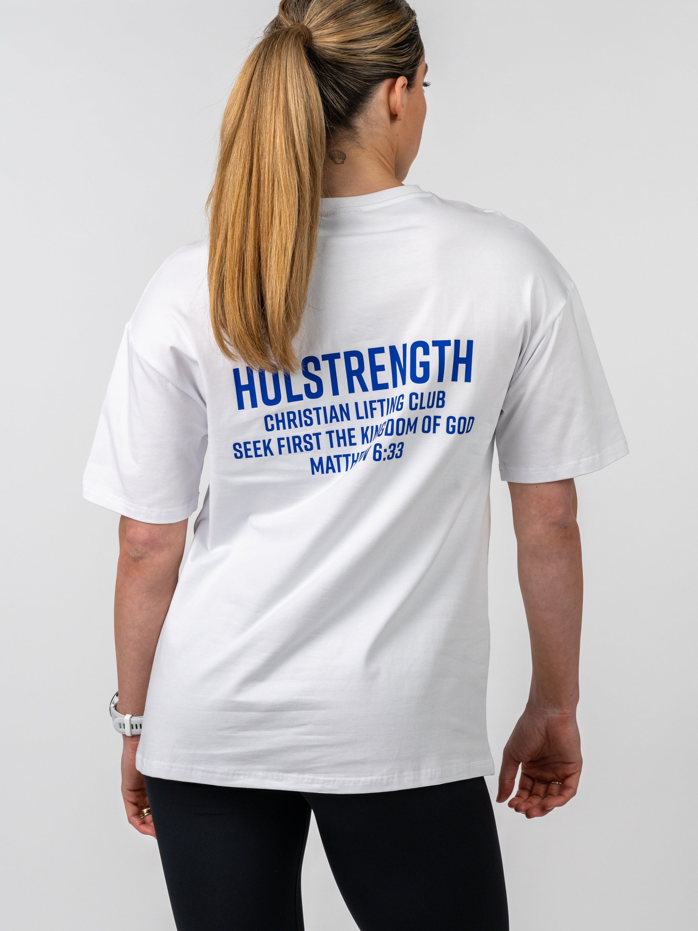 Woman seen from behind wearing a white Women's Lifting Club Oversized Tee with blue text referencing Christian lifting club and a Bible verse, paired with black leggings.