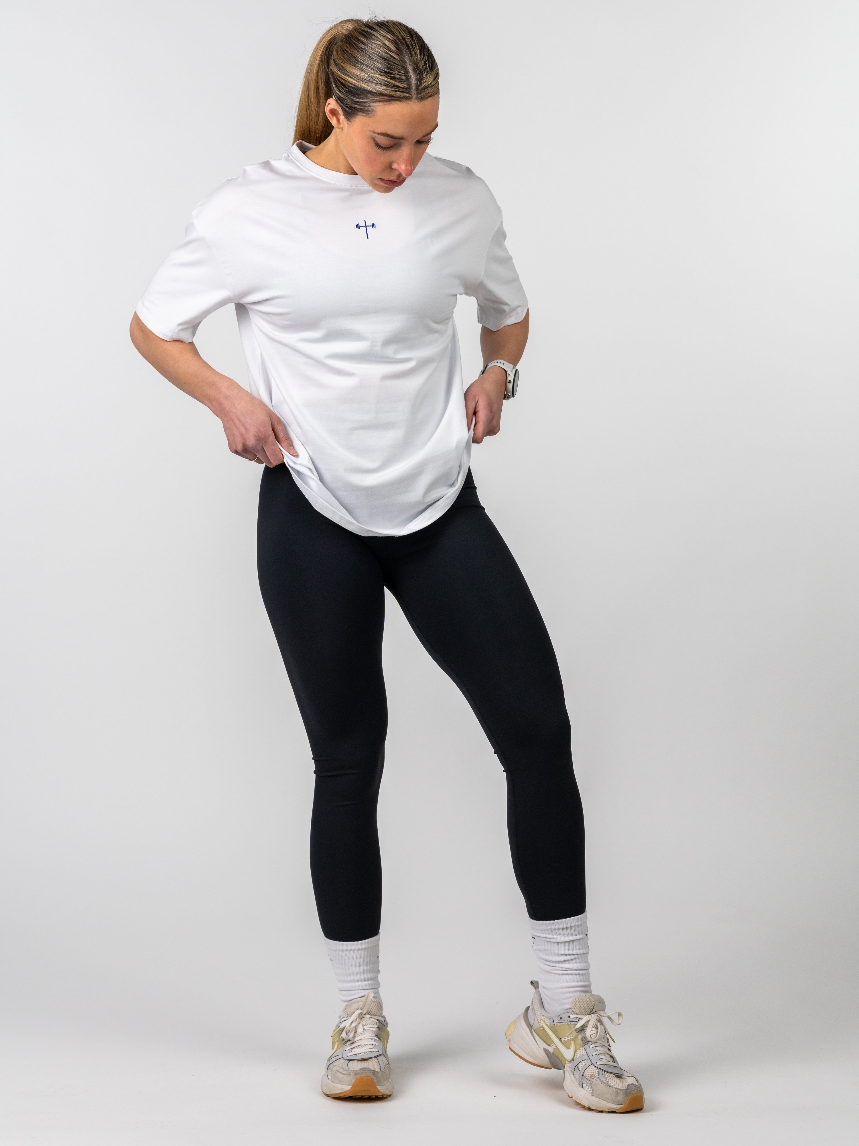 Woman wearing a white Women's Lifting Club Oversized Tee featuring a small blue cross and barbell graphic, paired with black leggings and sneakers in a studio setting.